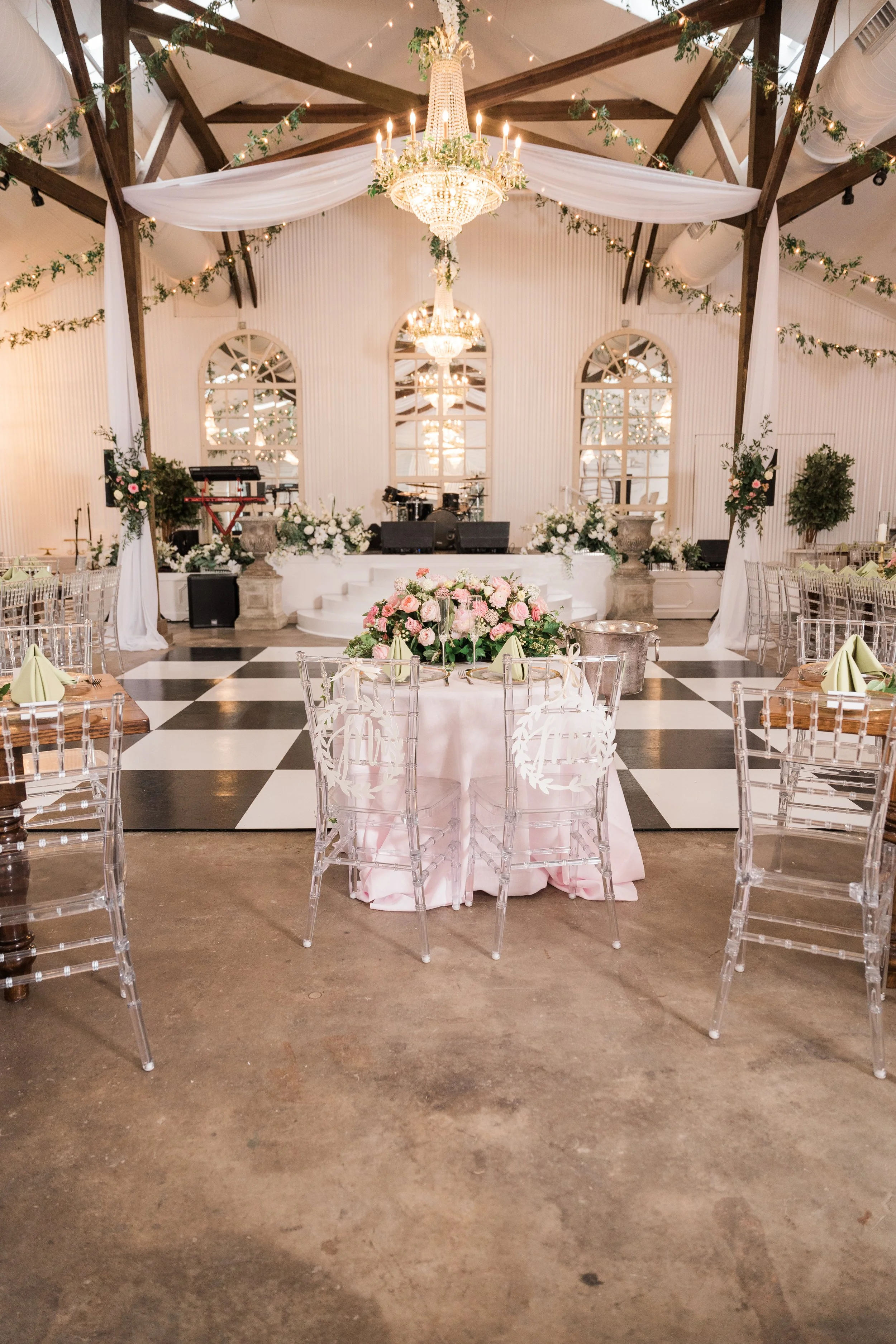 Elegant wedding reception setup with a checkered dance floor, round tables with pink linens and floral centerpieces, clear chairs, a stage with musical instruments, chandeliers, draped white fabric, string lights, and mirrors on the back wall.