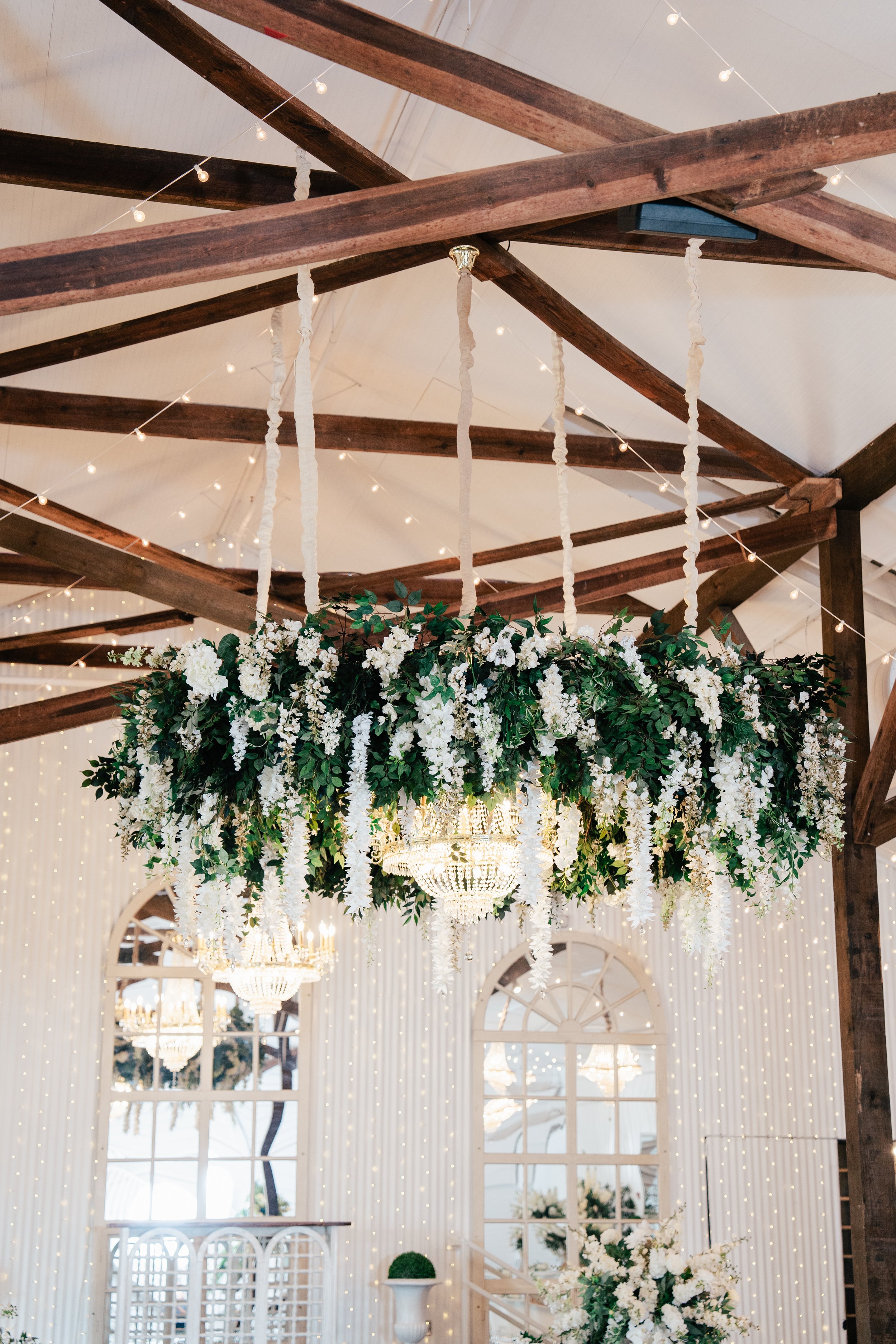 A large floral chandelier with white flowers and greenery hanging from a wooden ceiling with string lights in an elegant decorated event space.