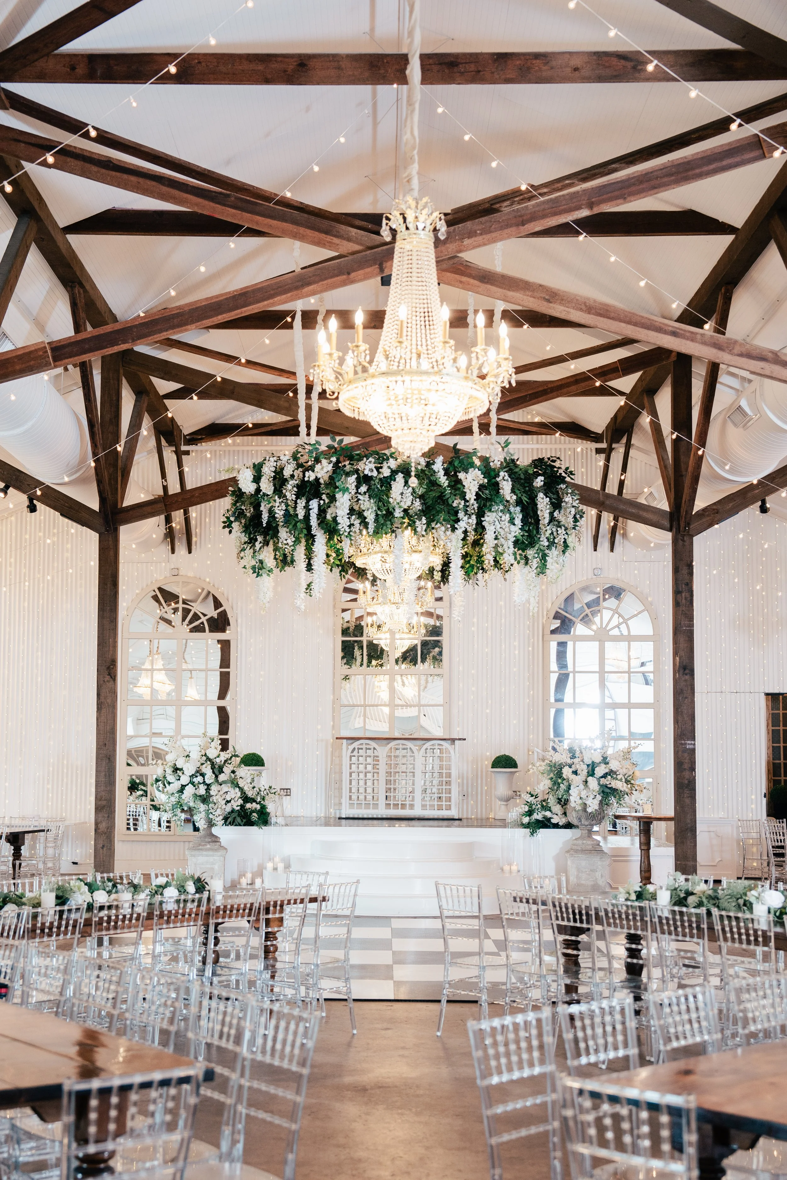 Elegant wedding ceremony venue decorated with white flowers and greenery, featuring large floral arrangements, a chandelier, string lights, and transparent chairs.