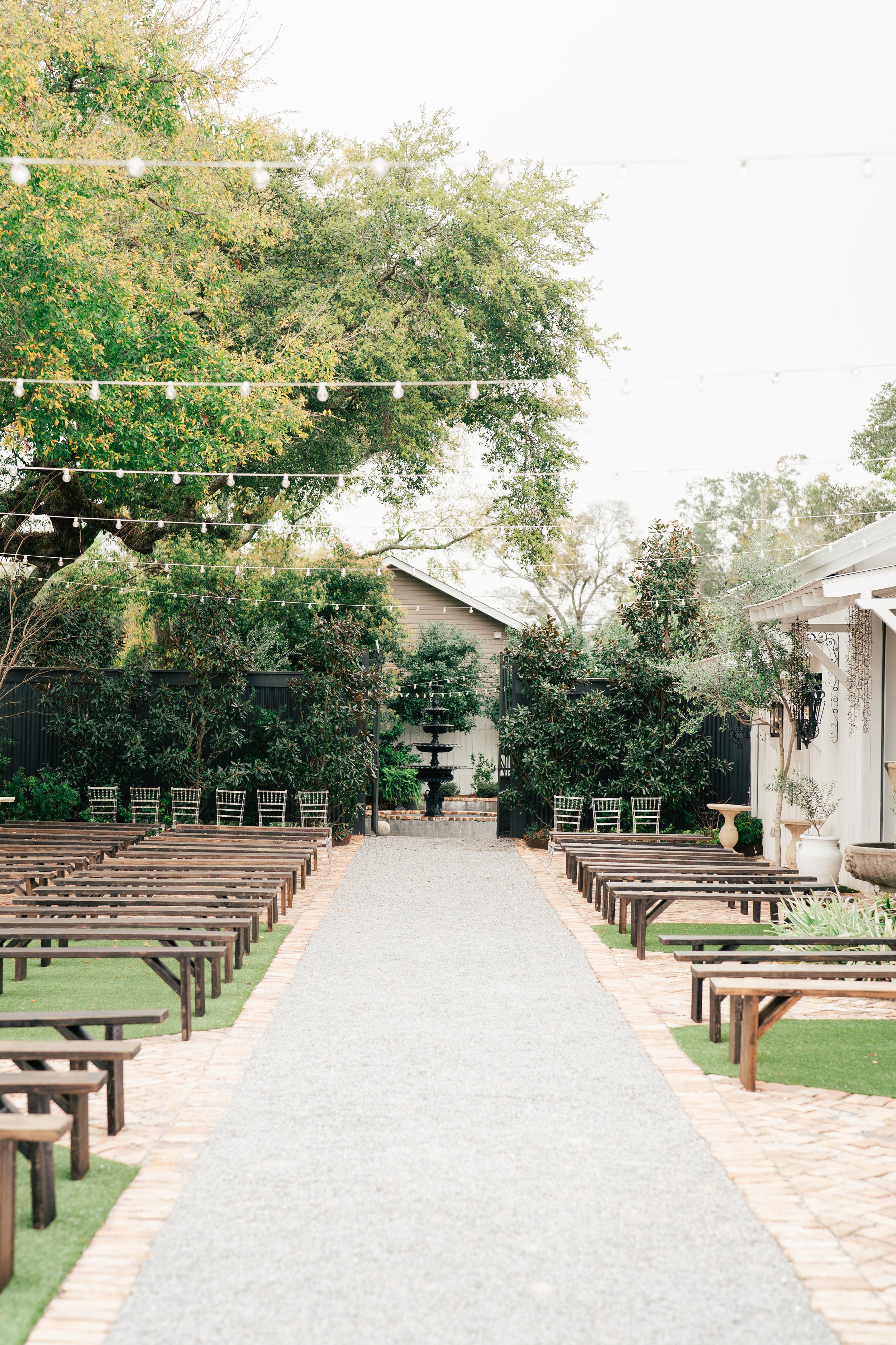 Outdoor Pensacola wedding setup with wooden benches on either side of a gravel aisle leading to a fountain, surrounded by trees, string lights, and a white building in the background.