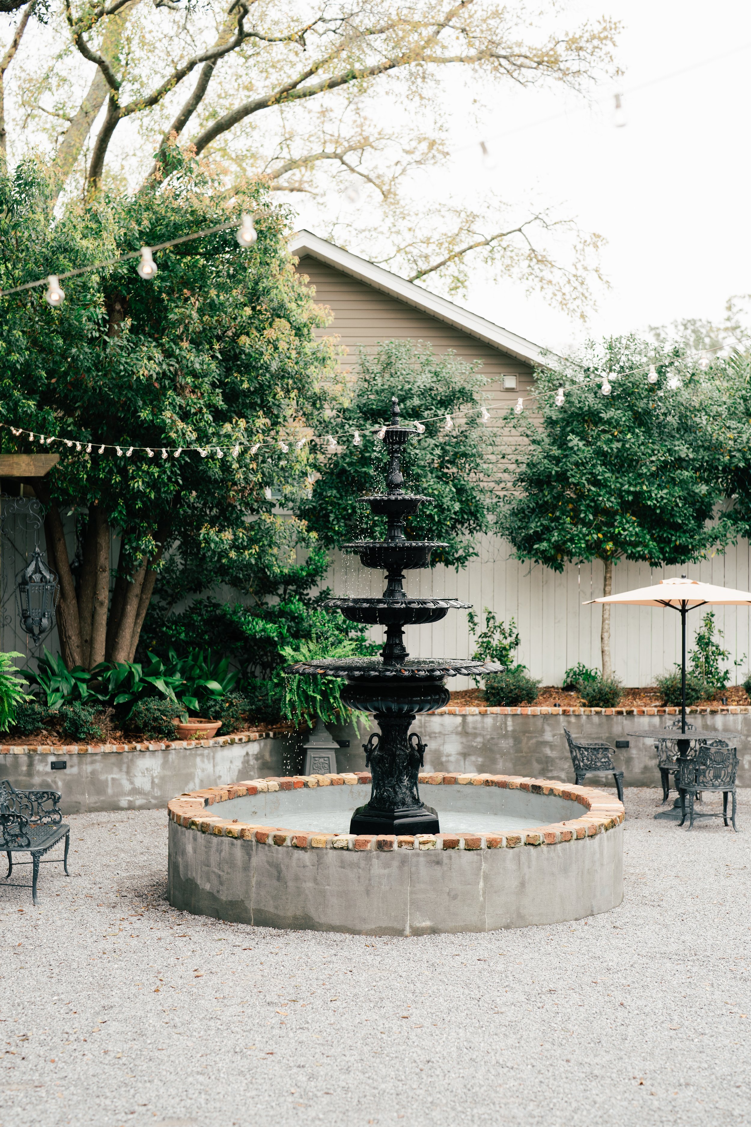 A decorative outdoor courtyard with a black tiered fountain in the center, surrounded by gravel, trees, and outdoor furniture, including a table with an umbrella and benches, and string lights overhead.