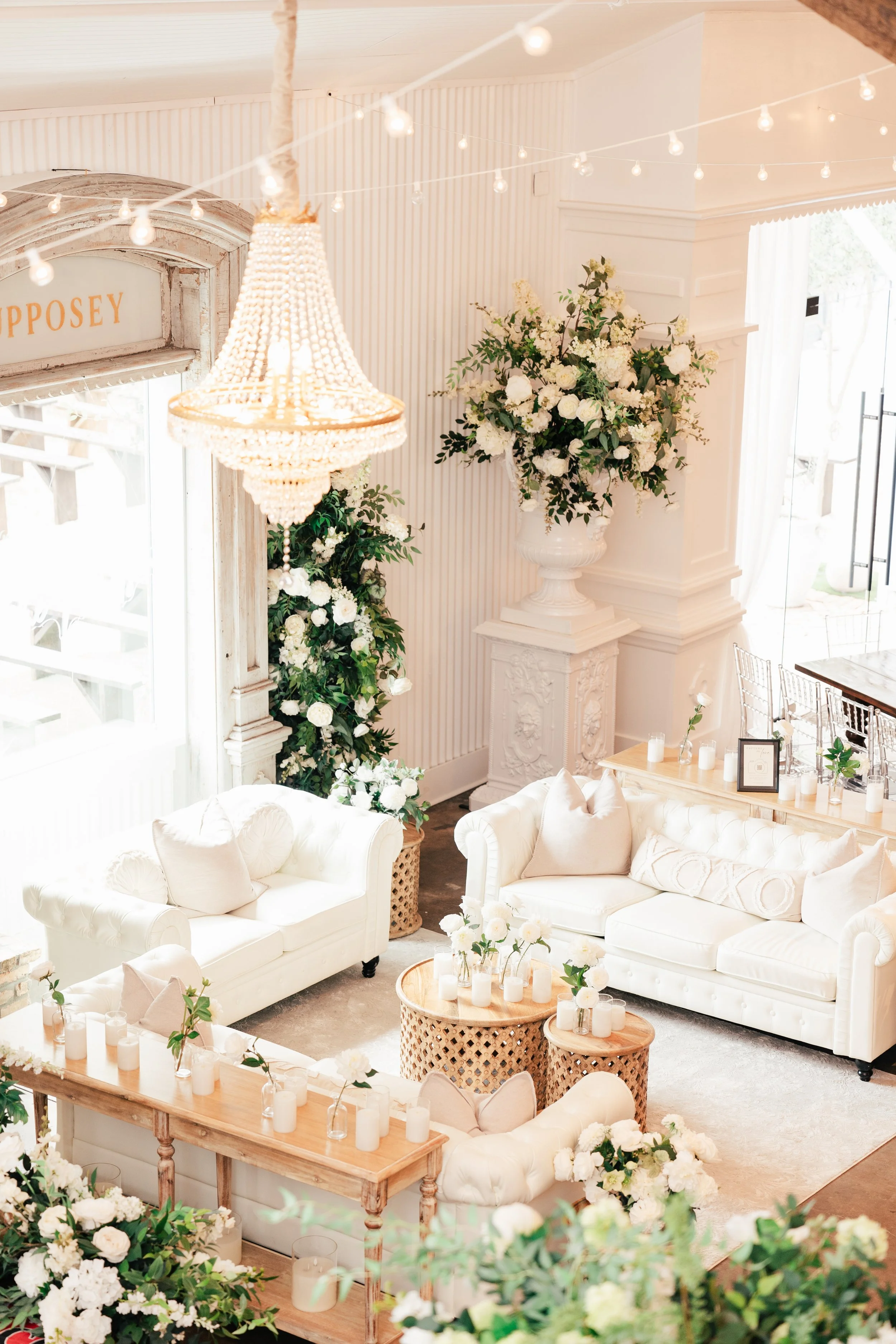 Elegant, bright room decorated with white sofas, floral arrangements, candles, and fairy lights, likely set for a wedding or special event.