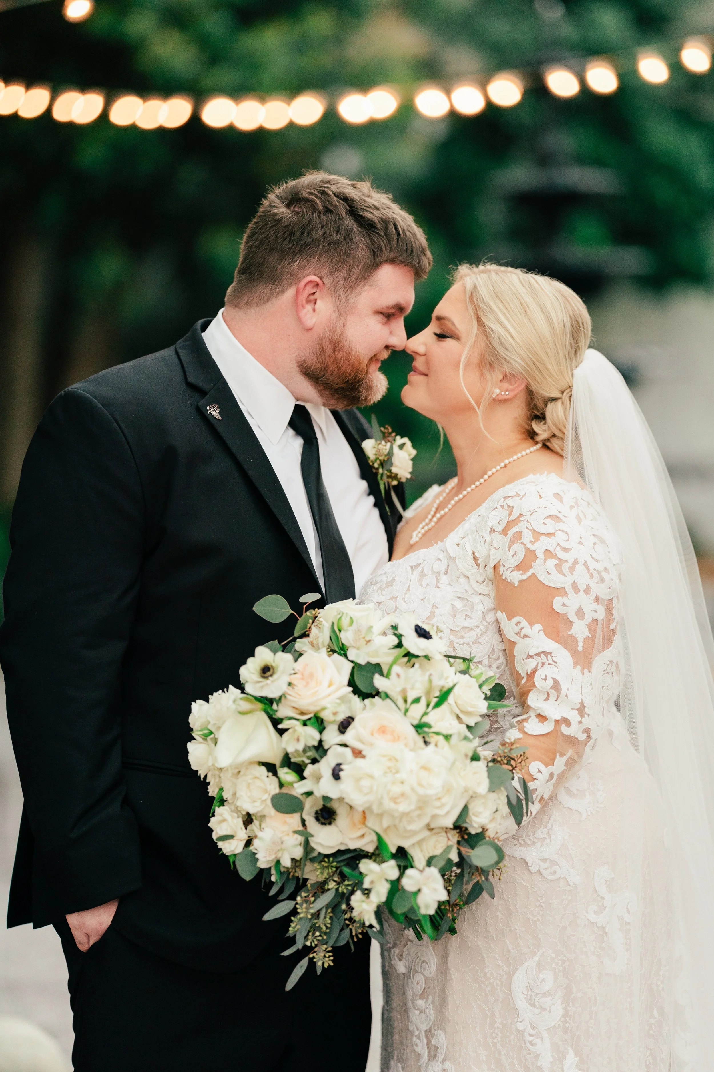 A Pensacola bride and groom on their wedding day, standing close with noses touching, outdoors with string lights in the background. The bride holds a large bouquet of white and blush flowers.