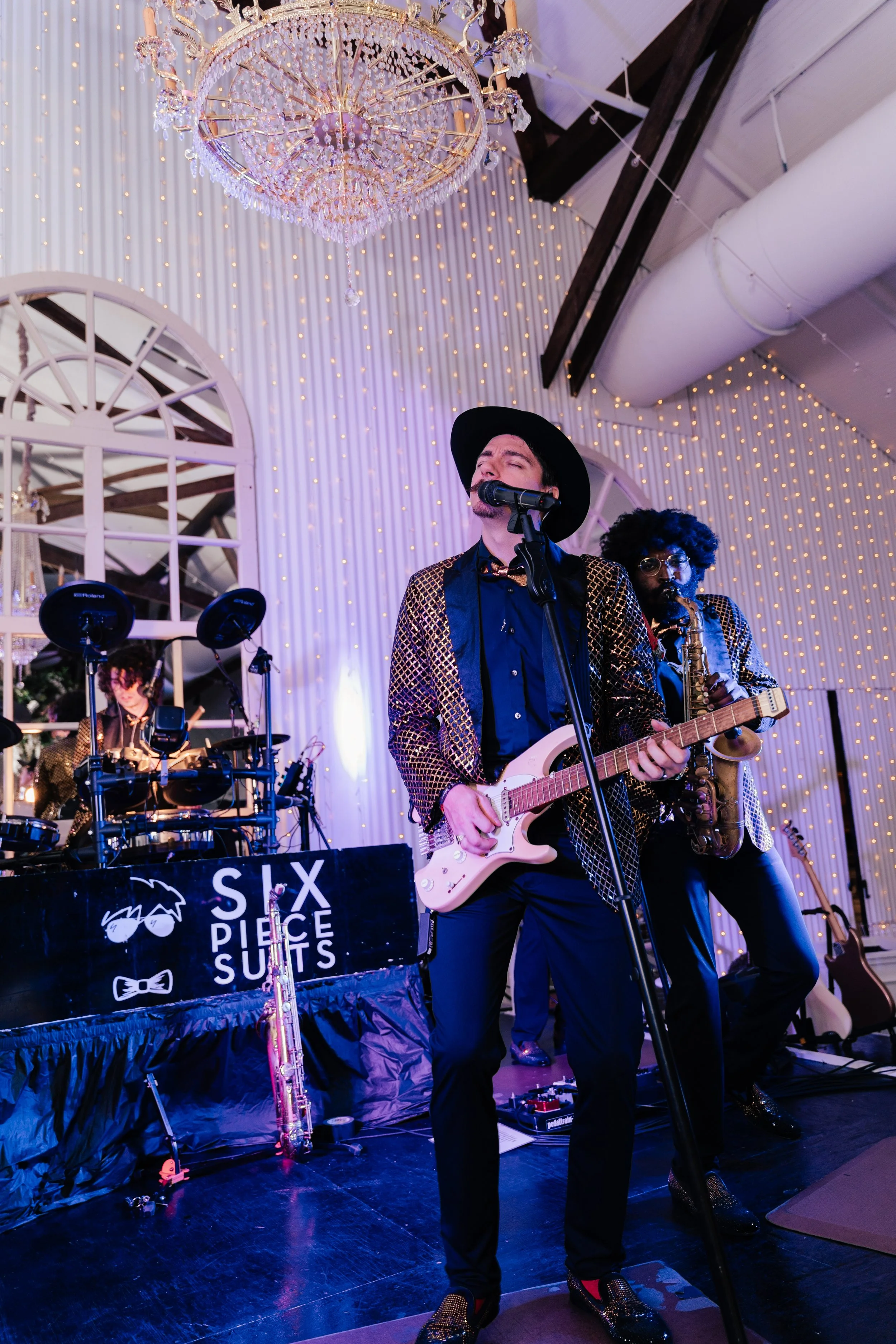 Musicians performing on stage at an event decorated with a chandelier, string lights, and elegant drapery. The lead singer is playing a pink electric guitar and wearing a black hat and glittery jacket. Additional band members play drums and saxophone
