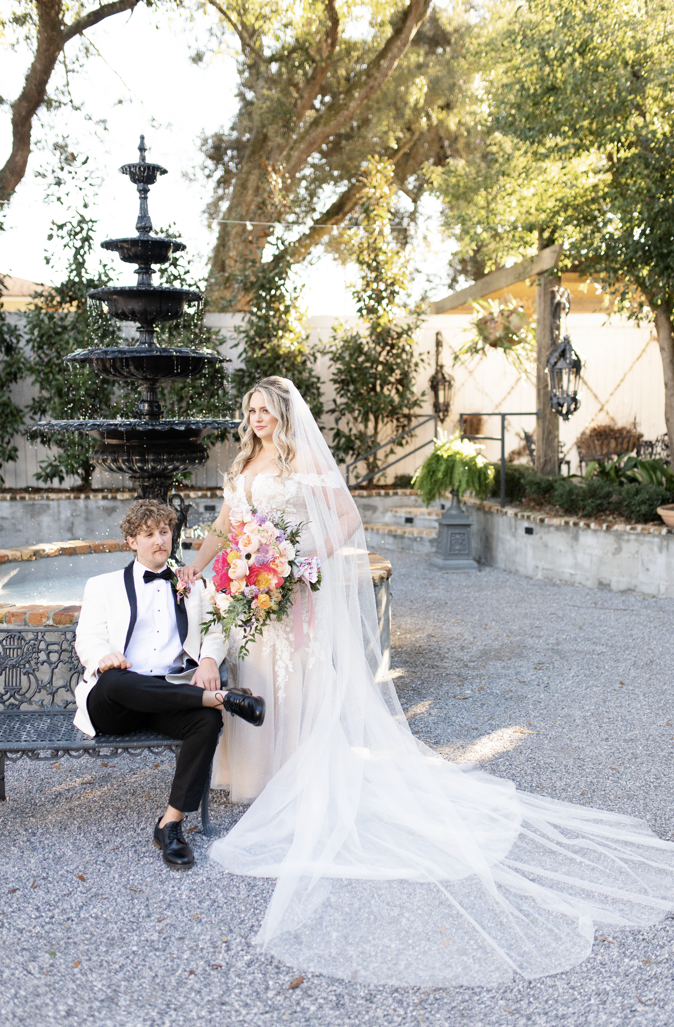 Bridal couple at a garden wedding, woman in a wedding dress and veil holding a bouquet, man in a tuxedo and bow tie sitting on a bench near a fountain.