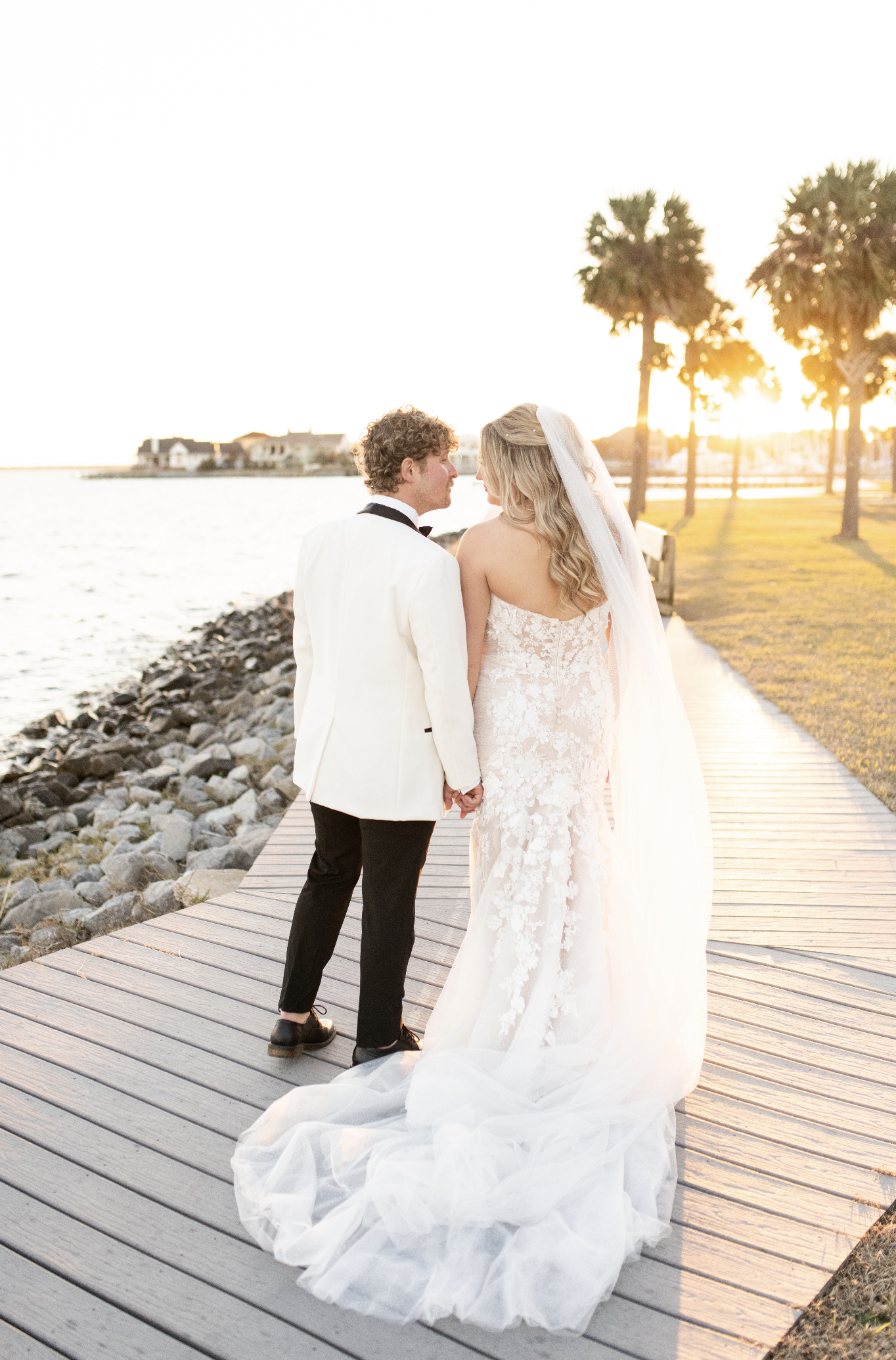 A Pensacola bride and groom holding hands on a wooden path by the water during sunset, with palm trees in the background.