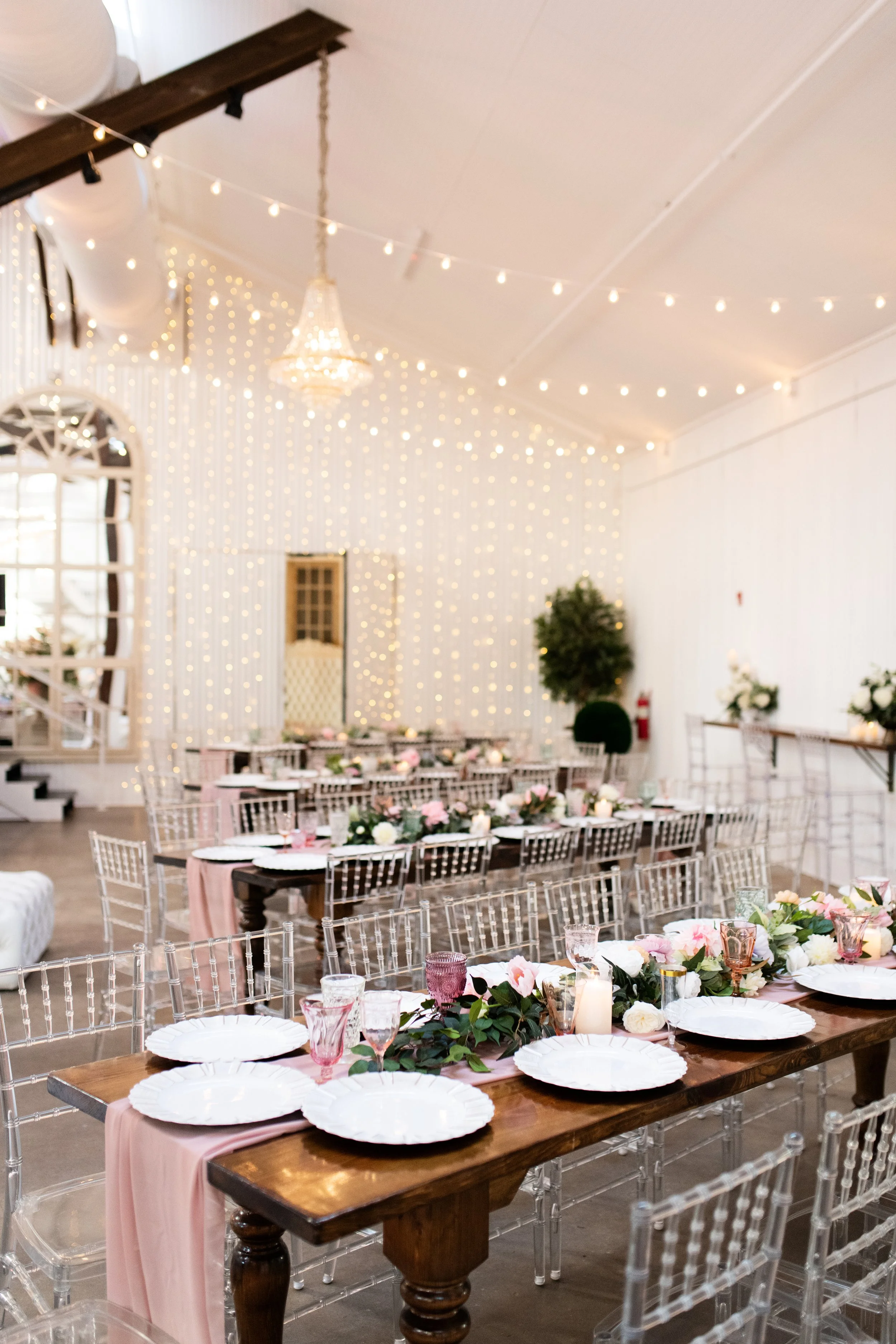 Elegant event space with long wooden tables, white plates, pink and white floral arrangements, candles, and transparent chairs, decorated with string lights and a chandelier.