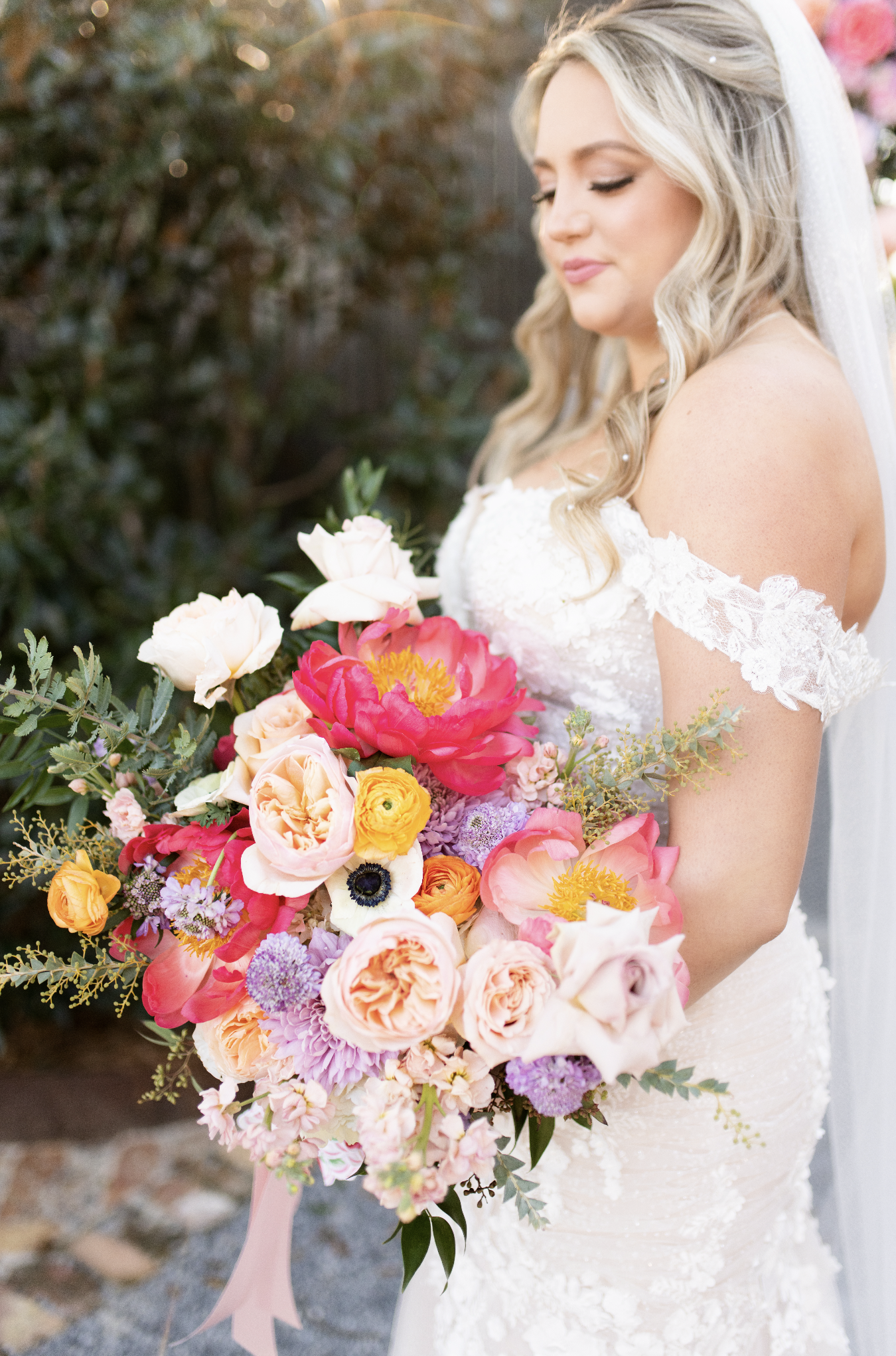 Brunette woman in wedding dress holding large colorful bouquet of flowers, outdoors with greenery