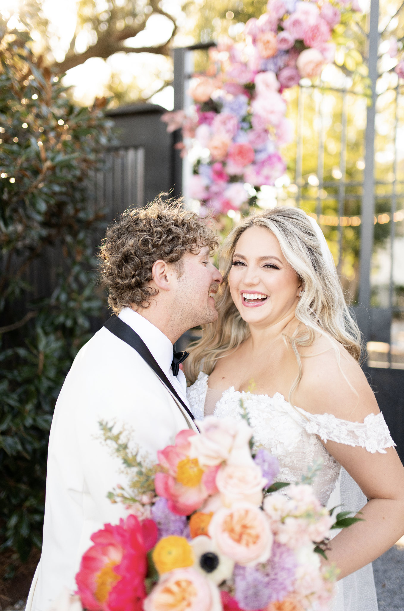 A new married couple smiling and laughing on their wedding day, with a colorful floral arrangement in the background and foreground.