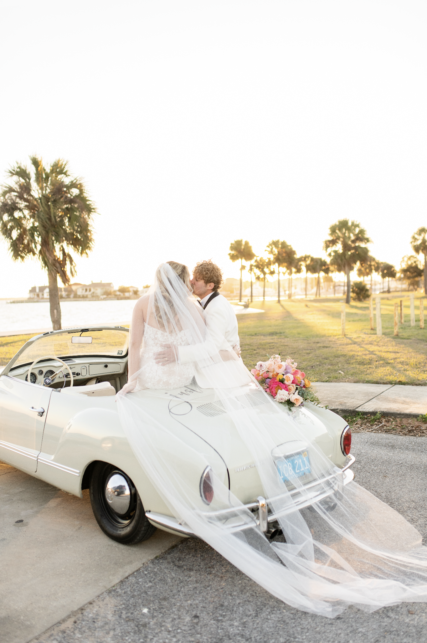 Bride and groom kissing in a vintage white convertible car decorated with a pink and white floral bouquet during sunset by a waterfront with palm trees.