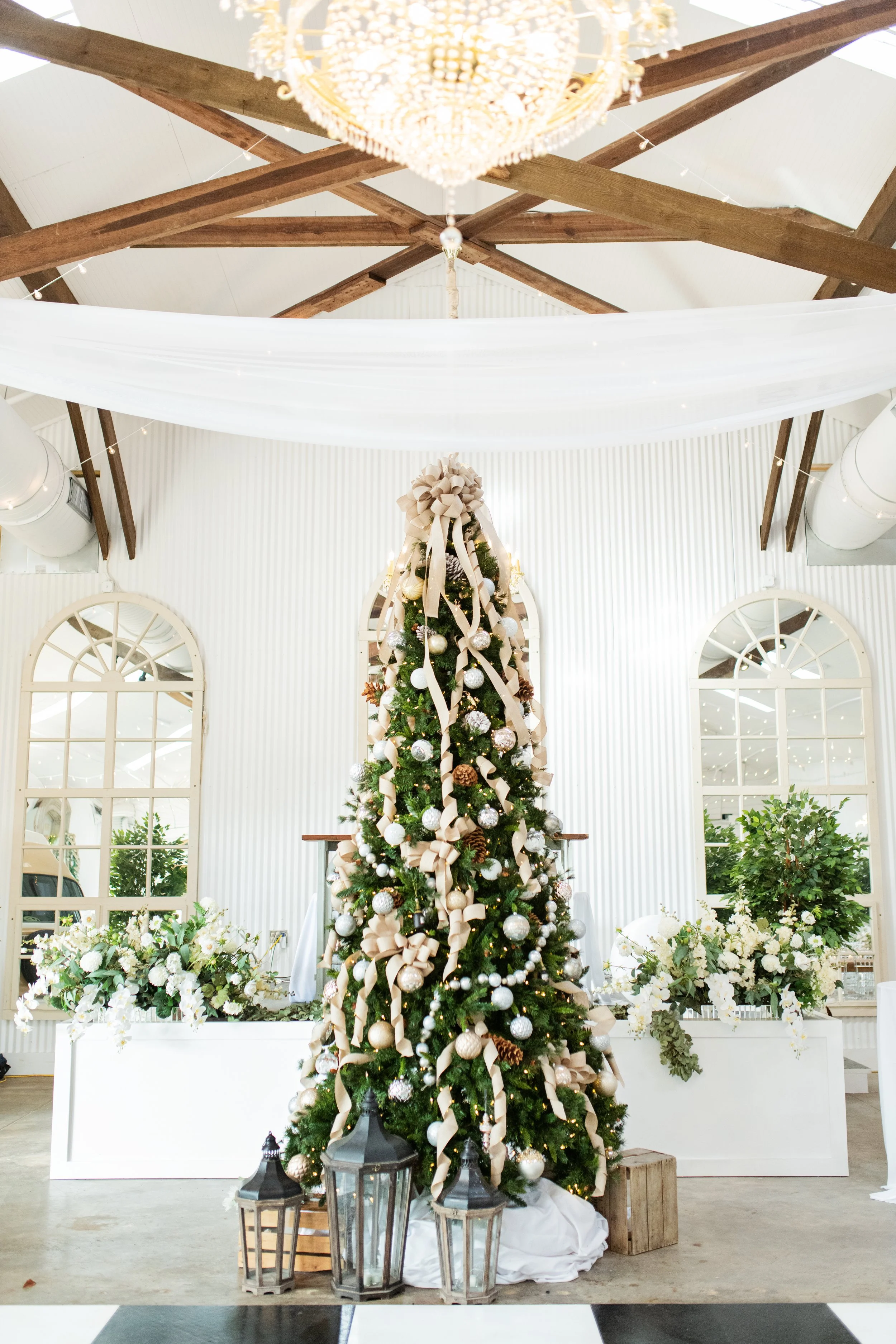 Decorated Christmas tree with white and gold ornaments, ribbons, and lights, topped with a bow, in a festive decorated room with white flowers and a chandelier in a Pensacola Wedding Venue.