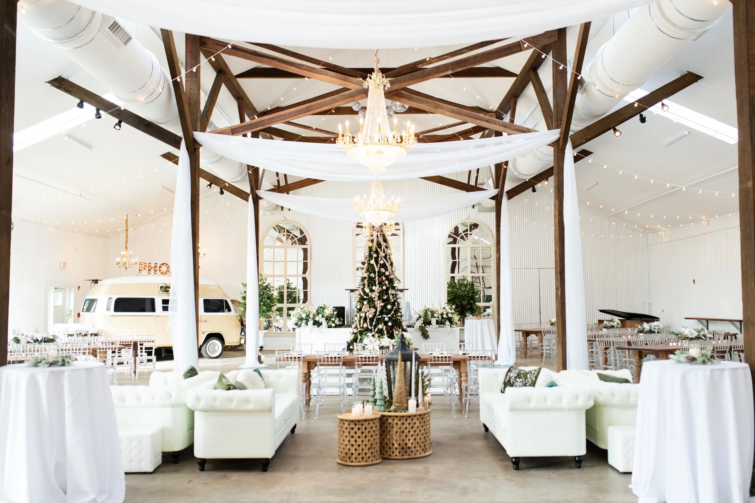Decorated Pensacola event space with a Christmas tree, vintage camper, chandeliers, white sofas, tables with candles, and string lights.
