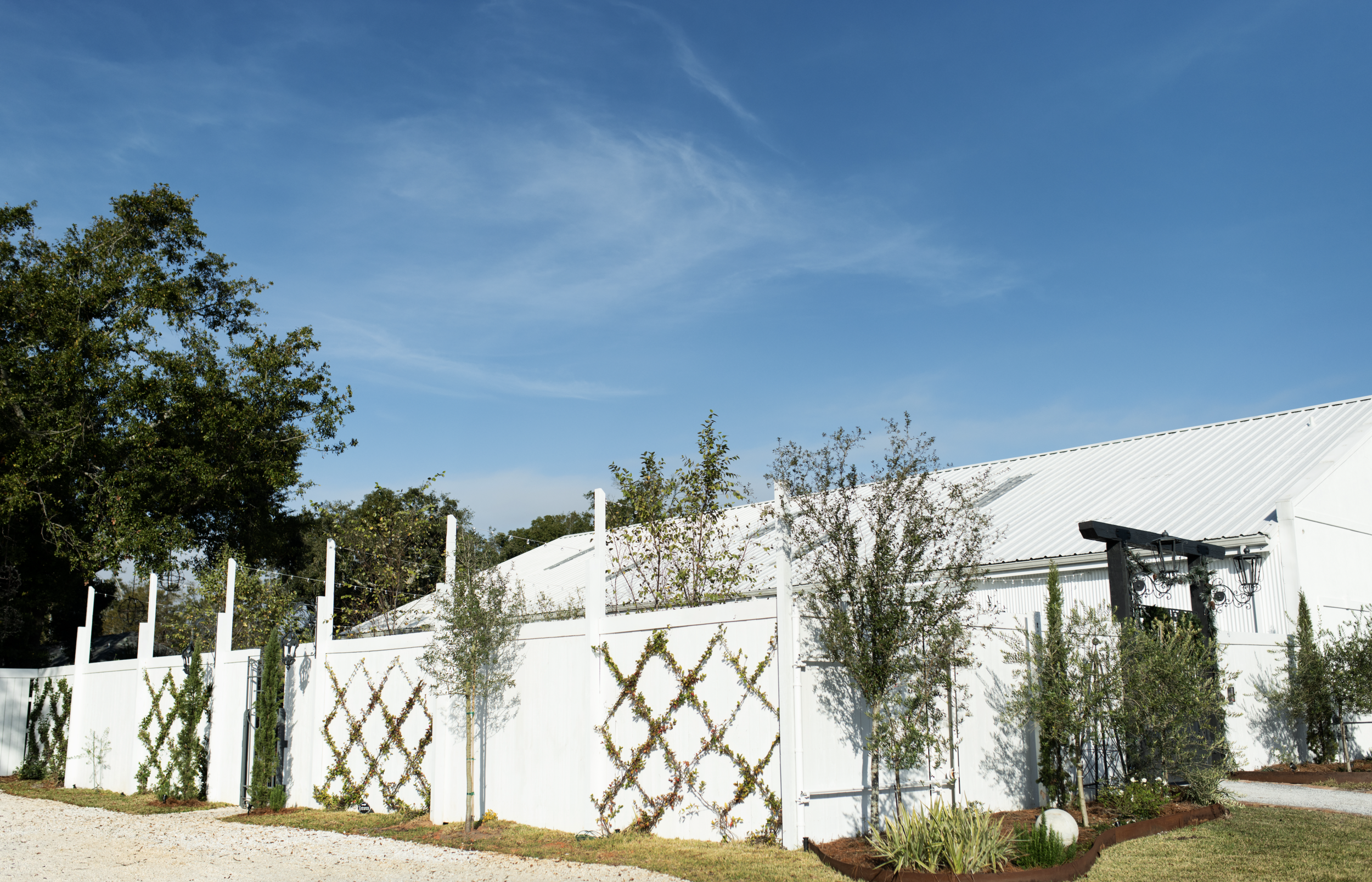 White fence with lattice decorations and small trees, a large white Pensacola Wedding Venue with a metal roof, and a clear blue sky.