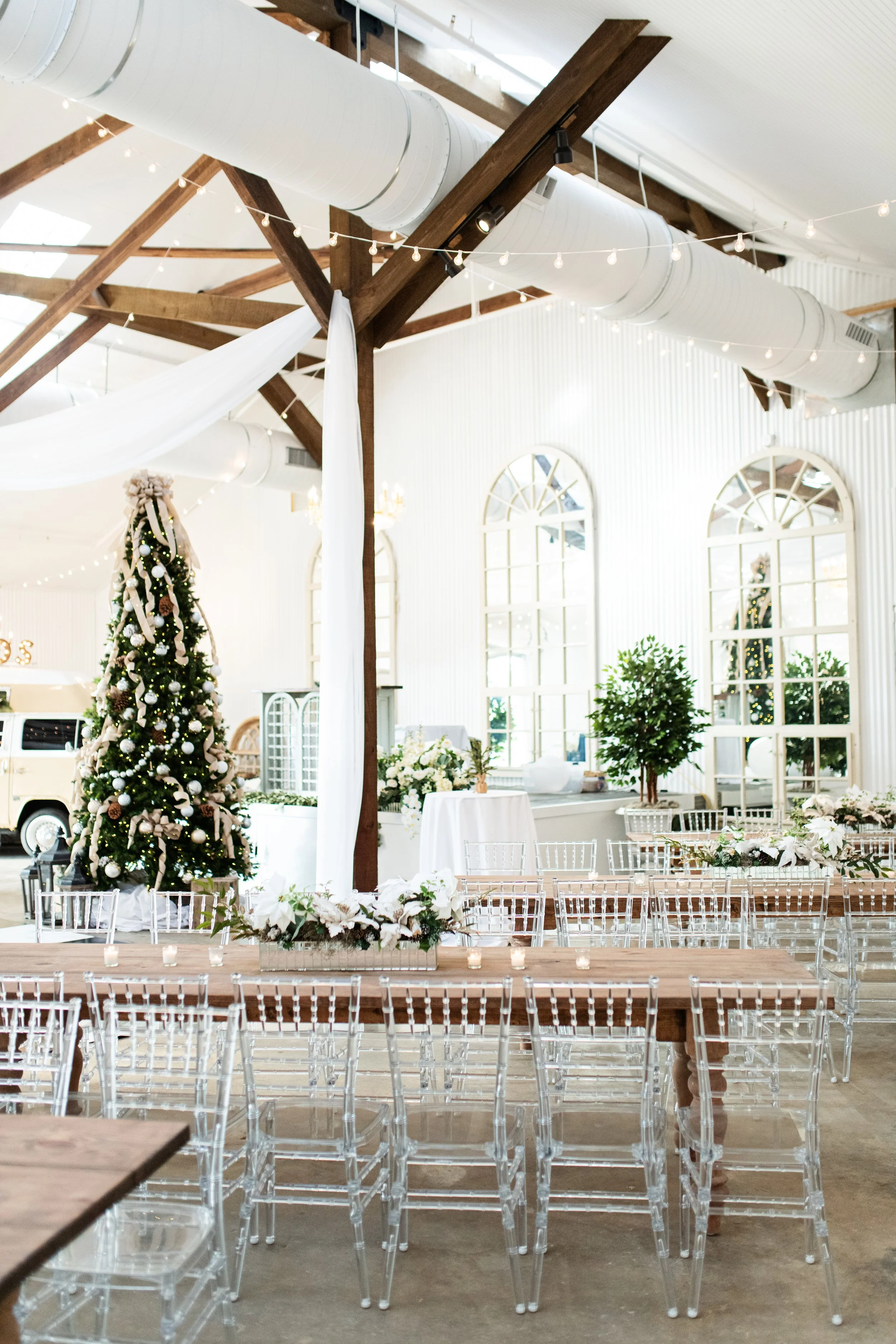 Decorated Christmas tree in a bright, airy venue with white walls, large arched windows, wooden tables, and transparent chairs, featuring string lights and holiday floral arrangements.