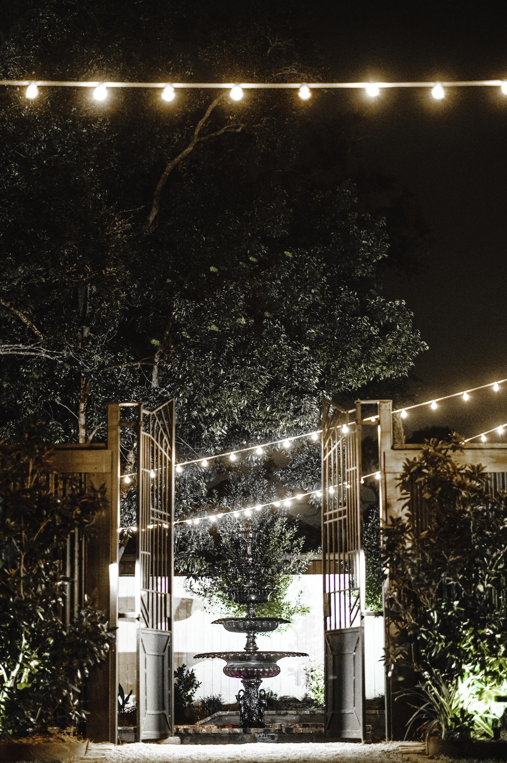 Nighttime outdoor scene with string lights, trees, an open gate, and a decorative fountain in the background in a Pensacola Wedding Venue.