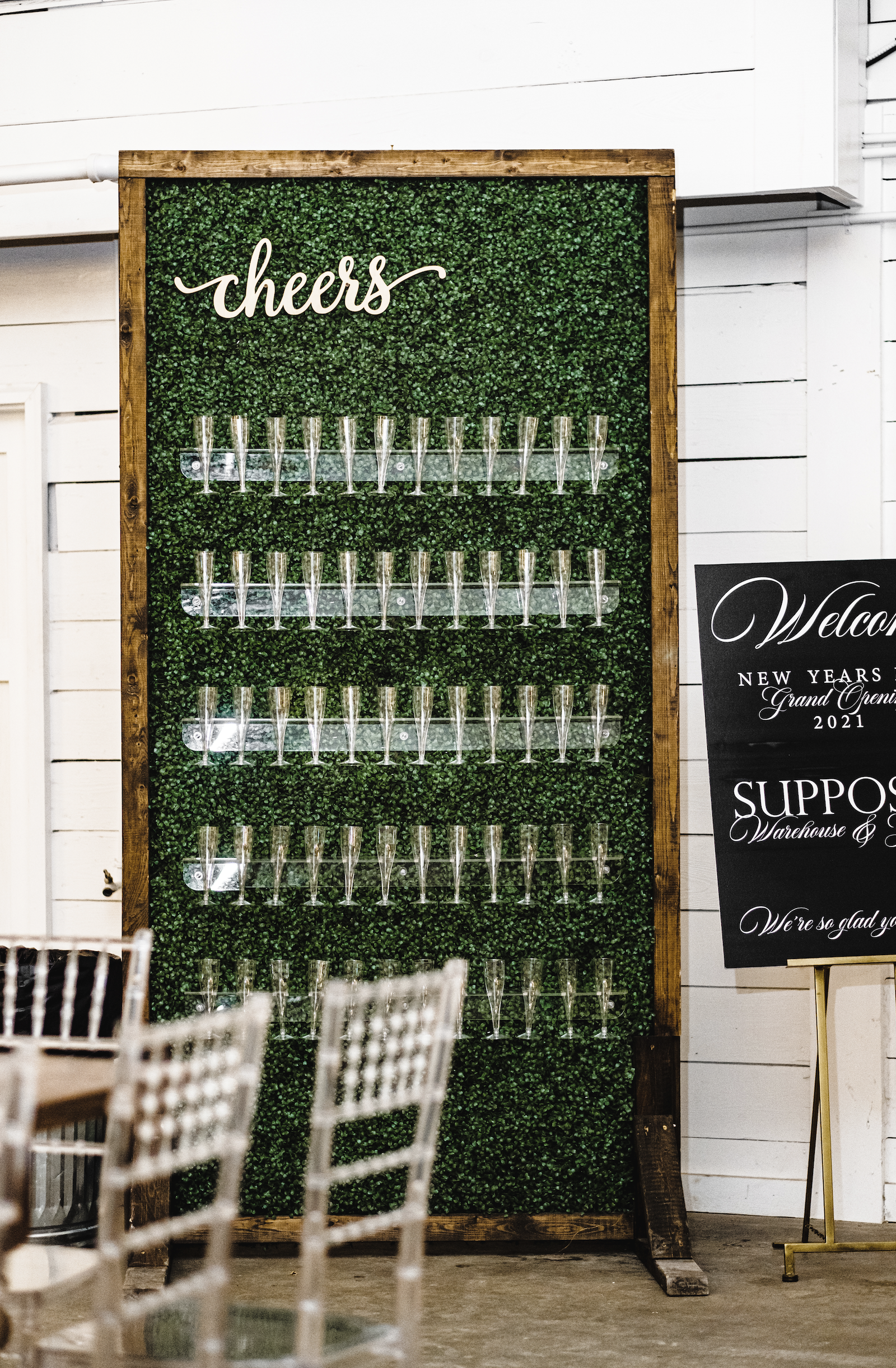 A decorative display with a green leafy background, the word "cheers" written in white script on top, and rows of champagne flutes attached to the backdrop, for a Pensacola Wedding.