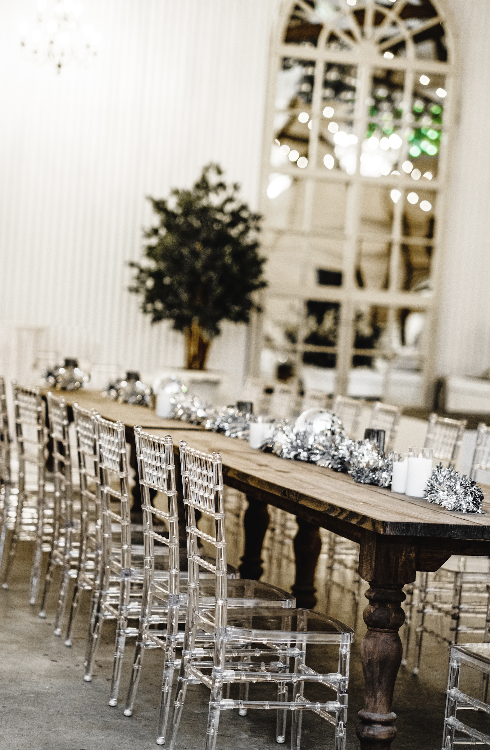 Decorated long wooden dining table with silver and white holiday centerpieces and clear chairs in a bright, elegant room with large mirrored wall and a tree in a Pensacola Wedding Venue.