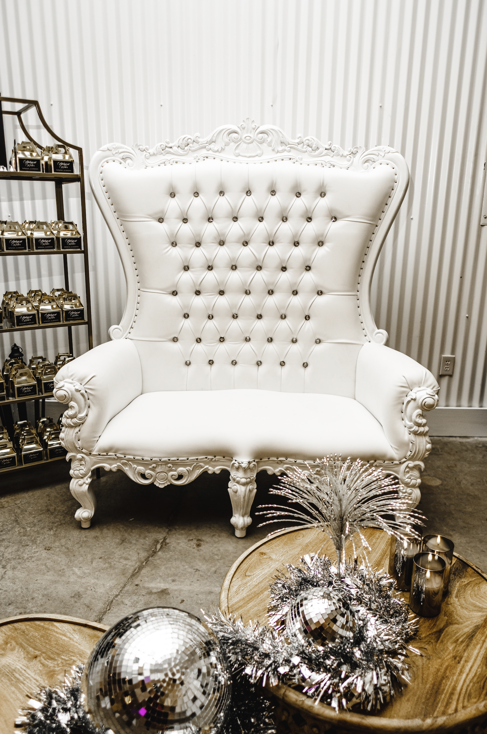 A white ornate high-back armchair with tufted upholstery and intricate carved wood details, set against a striped wall. To the left, a shelving unit displays multiple small gift boxes. In the foreground, a wooden table holds silver disco balls, tinse