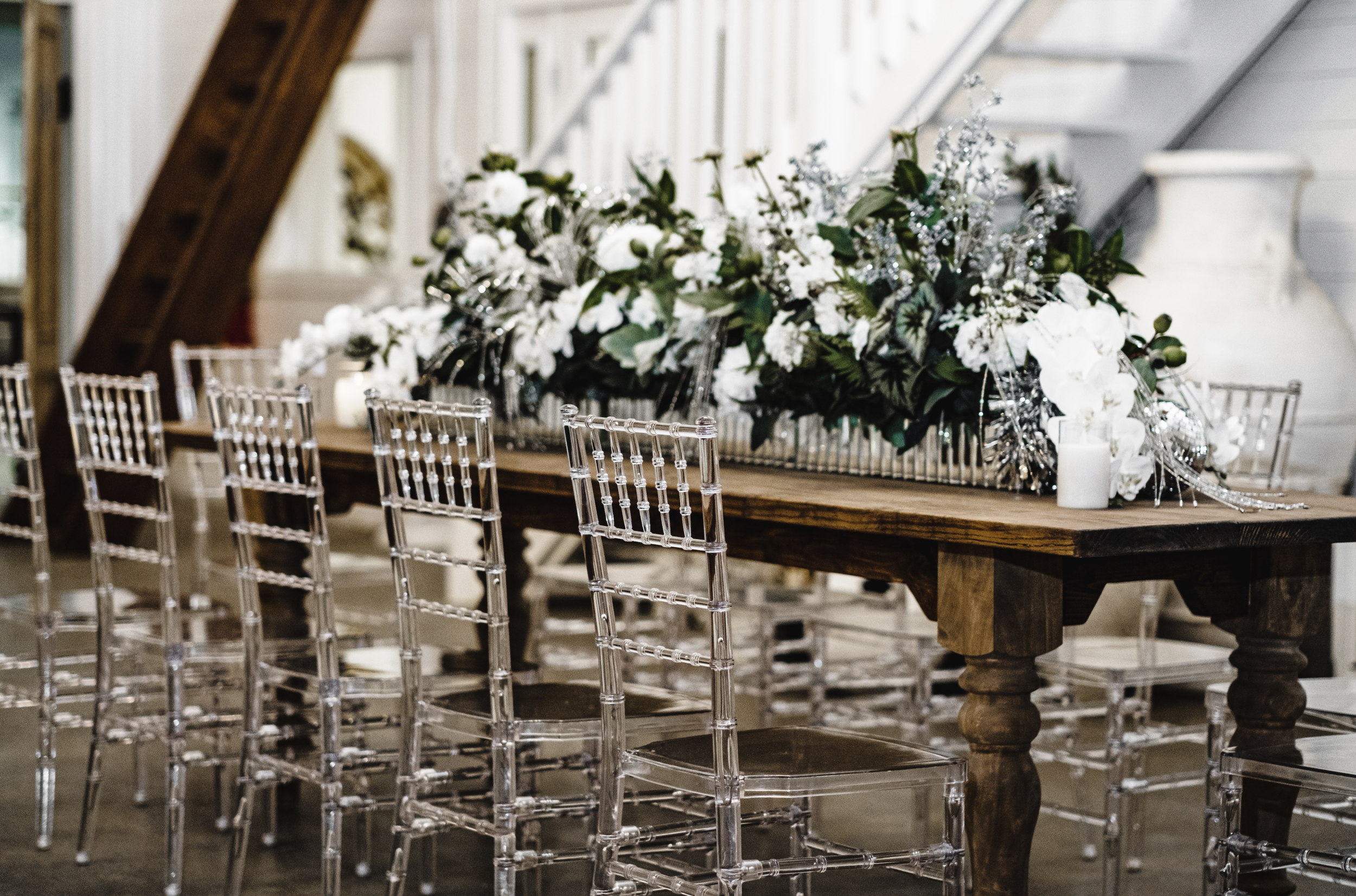 A long wooden table decorated with a large floral centerpiece of white flowers and greenery, surrounded by clear acrylic chairs, in an indoor setting with stairs in the background in a Pensacola Wedding Venue.