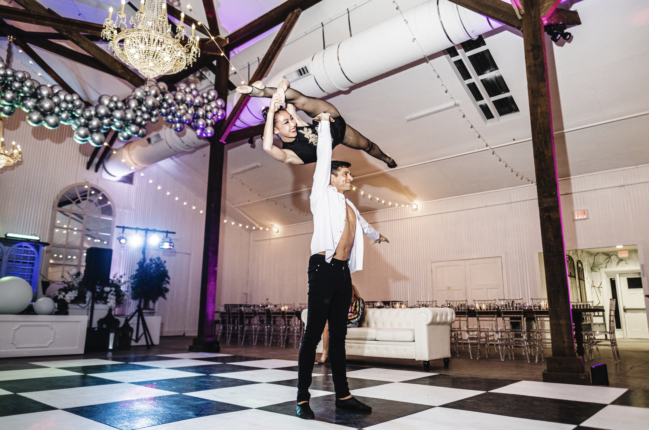 A male performer in black pants and a partially unbuttoned white shirt is holding a female performer above his head during a dance routine. The female performer is in a black dress and fishnet stockings, hanging upside down with her arm extended. The