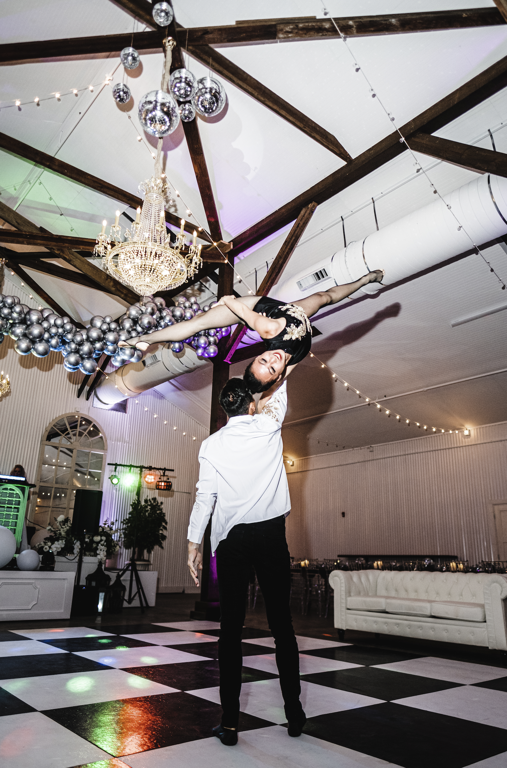 A couple dancing at an event with one person lifting their partner in a graceful pose inside a decorated Pensacola venue with chandeliers, balloon arrangements, and string lights.