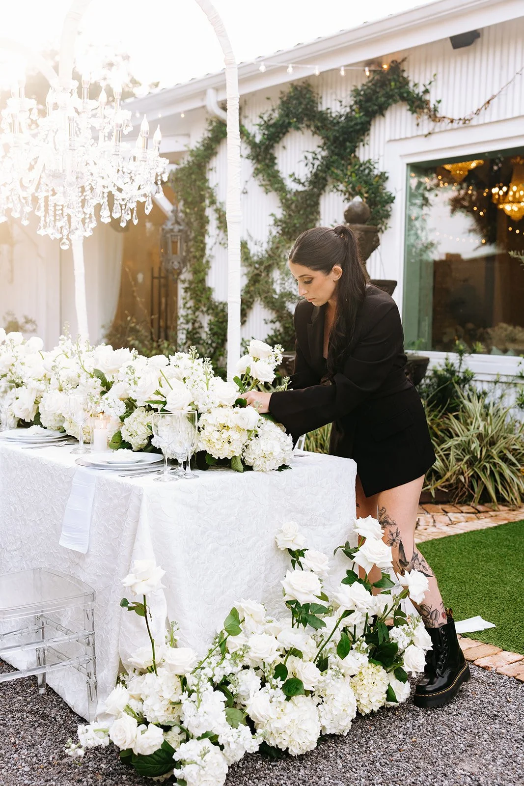 A pensacola Wedding venue owner arranging white floral centerpieces on a white banquet table outdoors, decorated with white roses, hydrangeas, candle holders, and a chandelier hanging above, at a garden event during sunset.