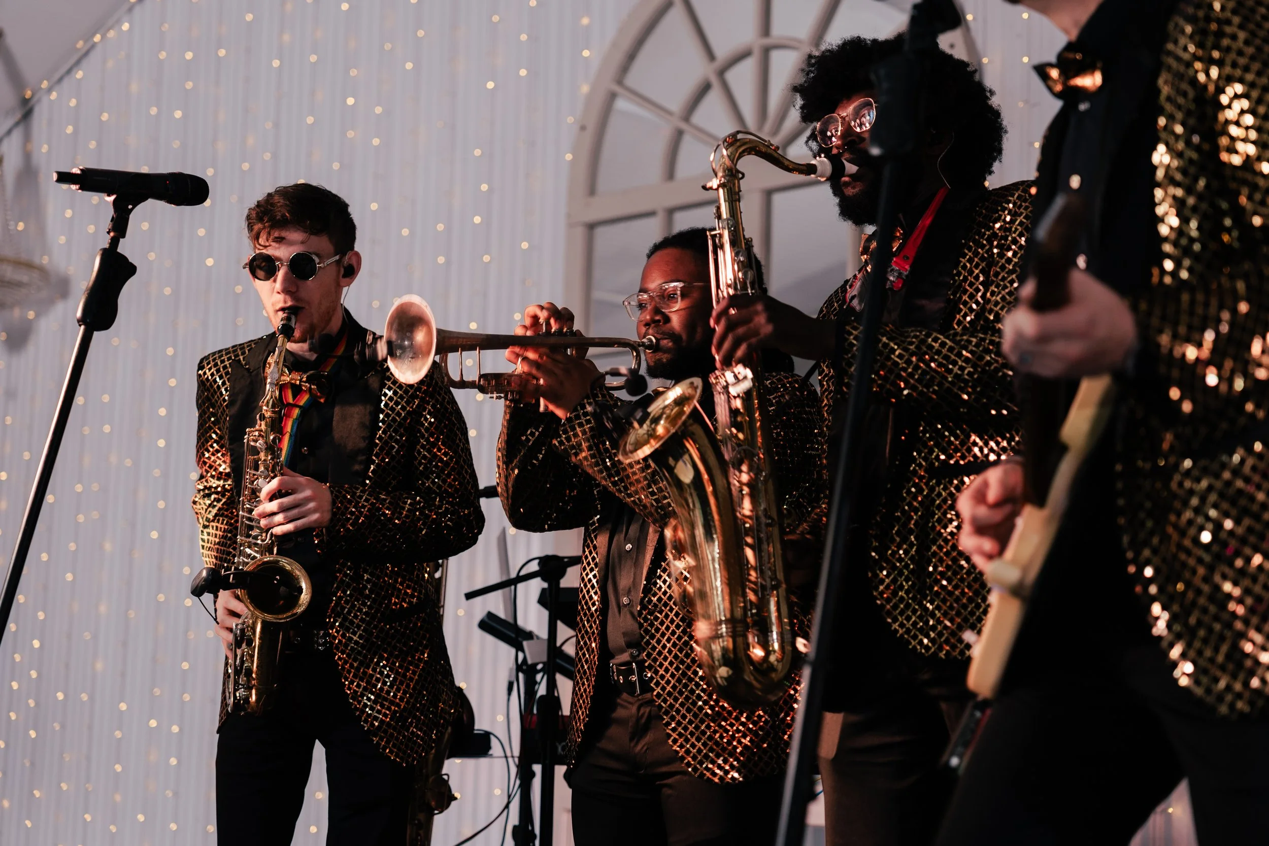 Jazz band performing on stage, musicians wearing gold sequin jackets, playing saxophone, trumpet, and guitar, with a microphone and string lights in the background in a Pensacola, Florida Wedding Venue.