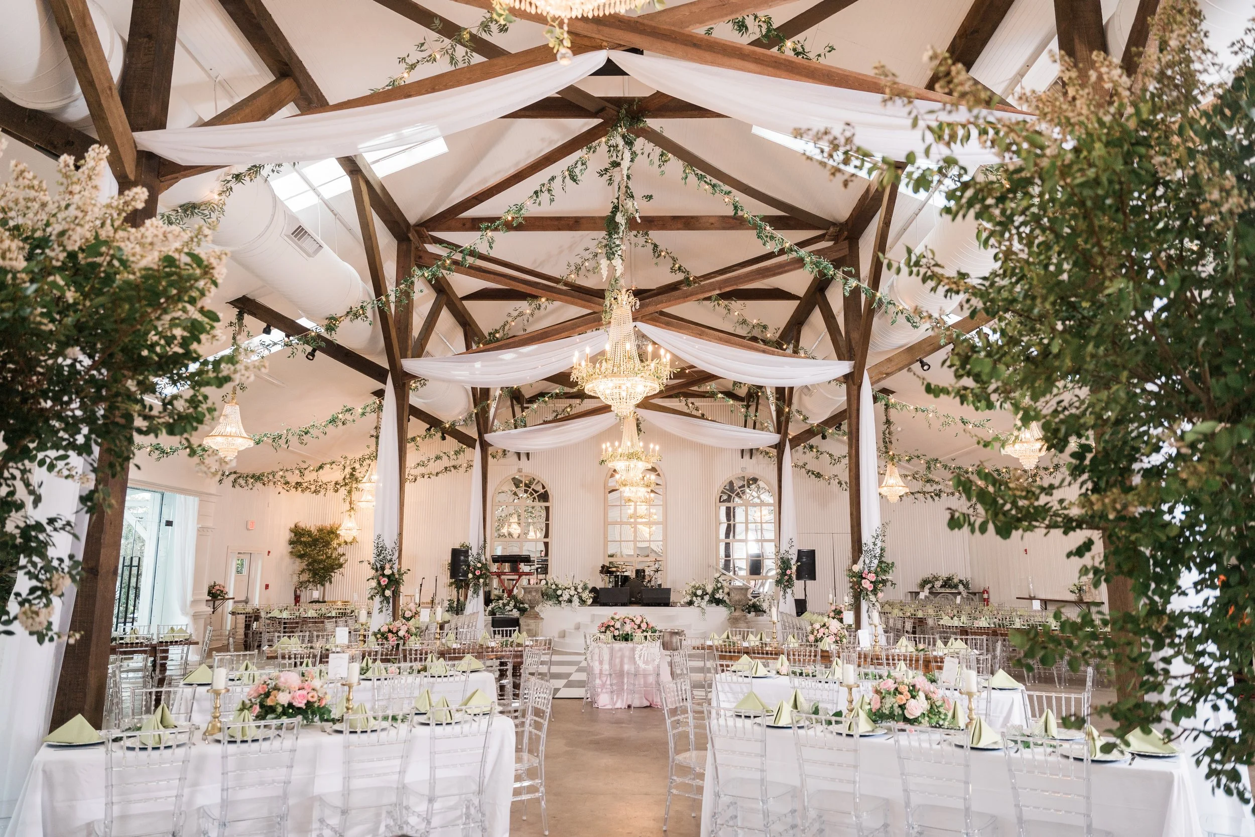 Elegant wedding reception setup in a spacious hall with draped white fabric and greenery on the ceiling, chandeliers, floral centerpieces, and white chairs around tables in Pensacola.