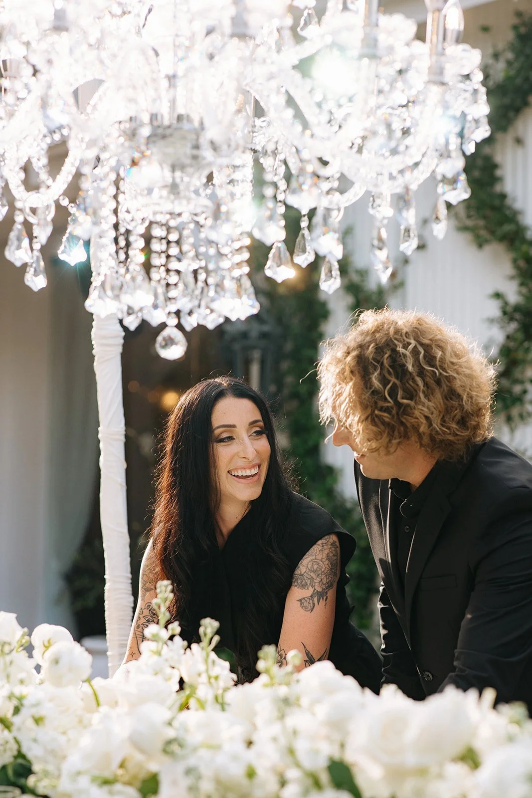 Two wedding venue owners sitting at a table with white flowers, smiling and talking, underneath a crystal chandelier outside in daylight.