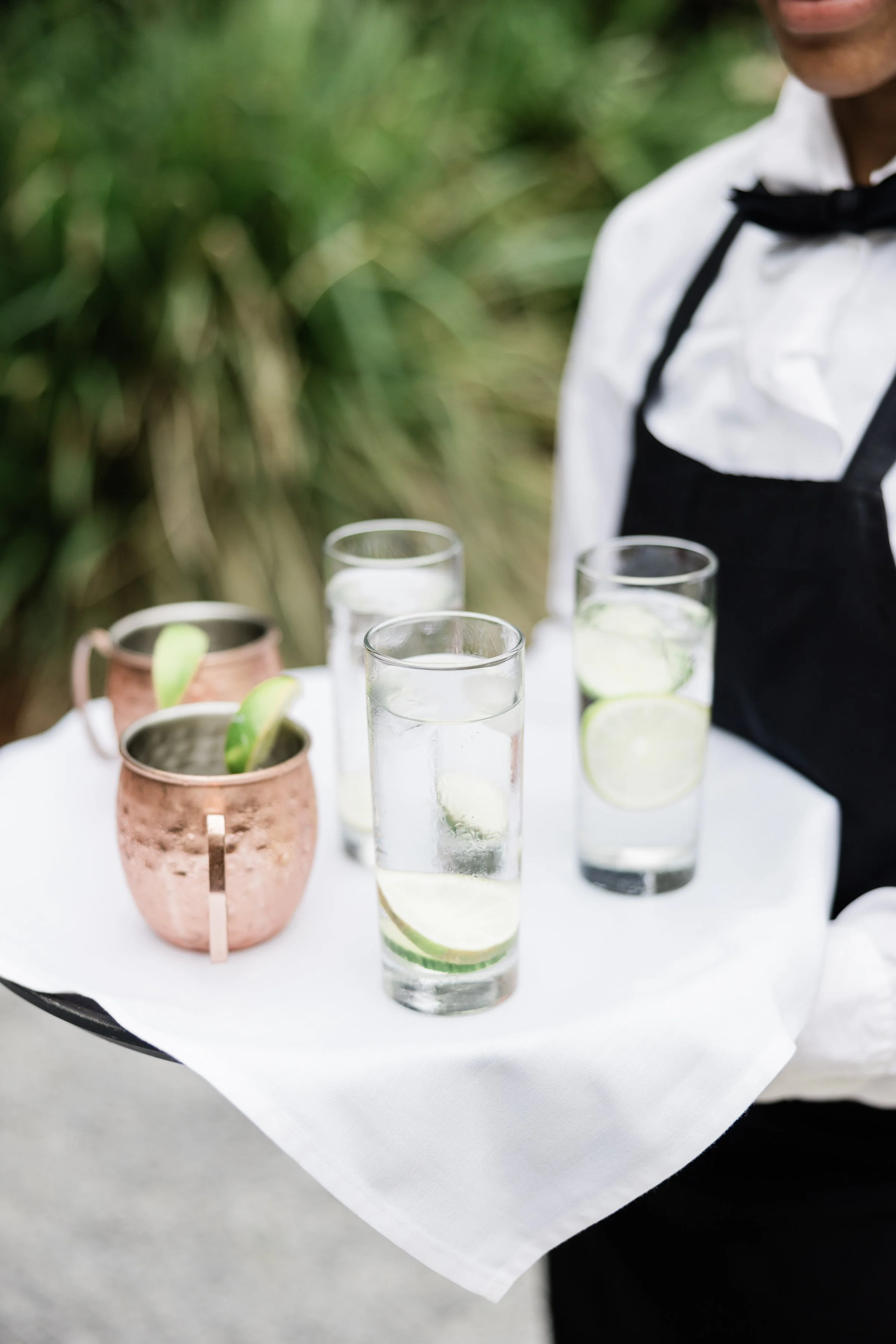 A server holding a tray with four glasses of water and slices of lime, two copper mugs with lime, against a green outdoor background in a Pensacola Wedding Venue