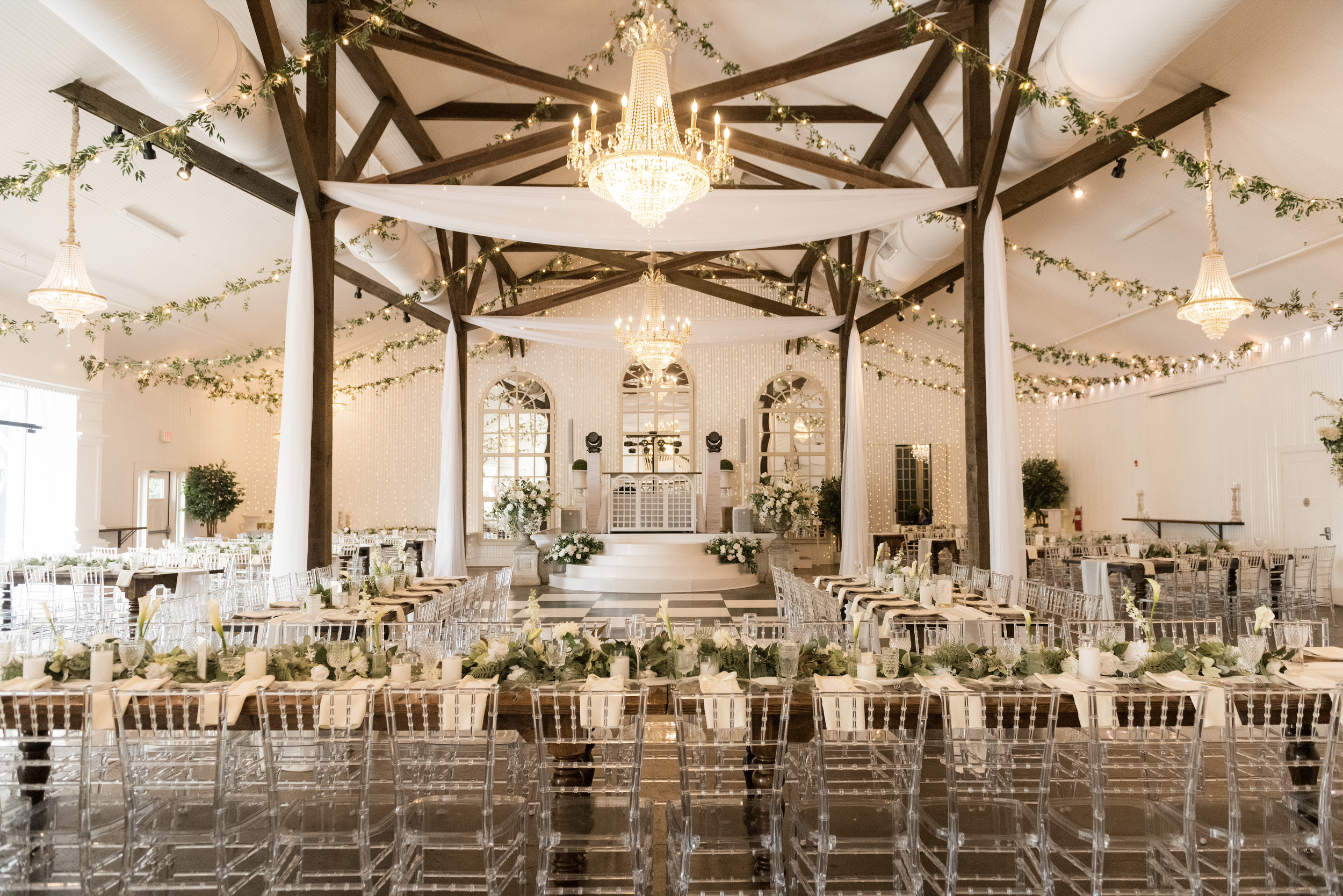 Elegant wedding venue reception hall decorated with white drapes, greenery, string lights, and chandeliers, with long tables set with candles and floral arrangements, and a dance floor illuminated by chandeliers at the front in Pensacola.