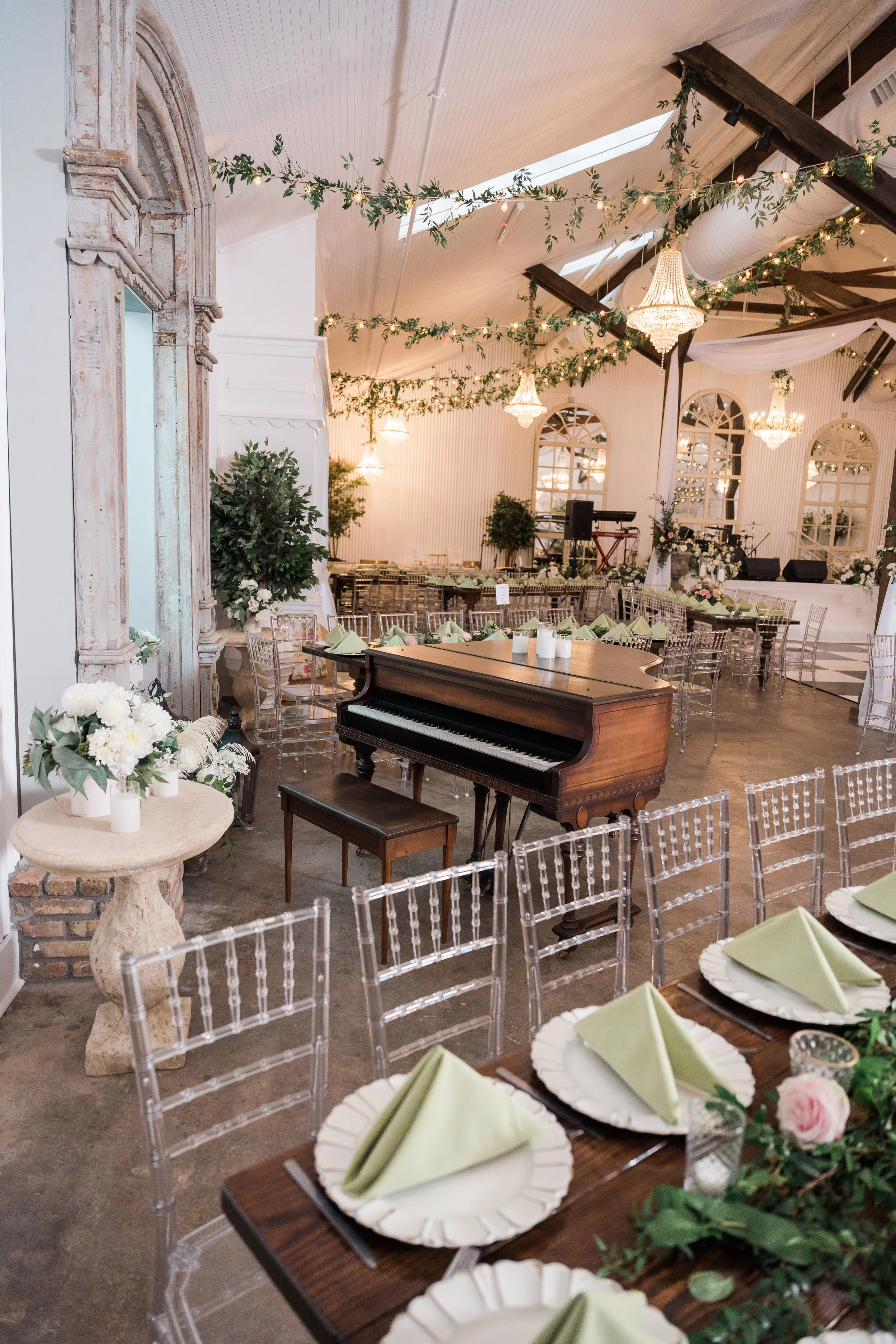 Elegant wedding and event banquet hall decorated with greenery and chandeliers, featuring a grand piano, set tables with plates and folded napkins, and floral arrangements for a celebration or wedding.