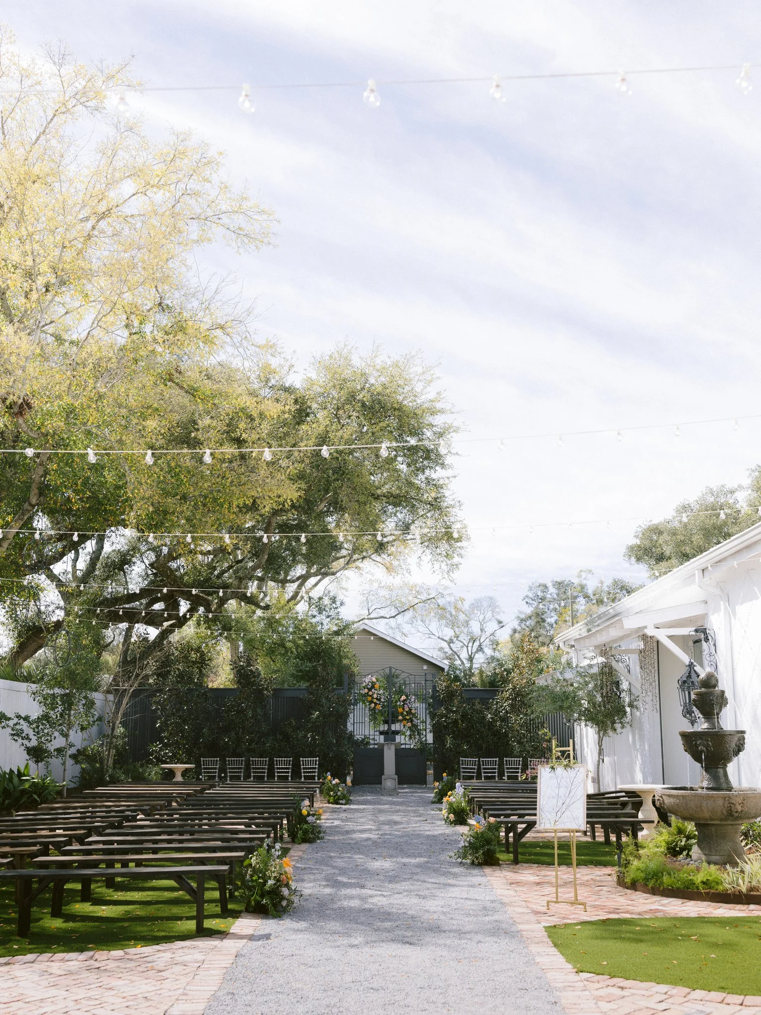 Outdoor wedding venue in Pensacola with rows of benches, string lights, and a decorated altar at the end of the aisle, surrounded by trees and a white building on the right.