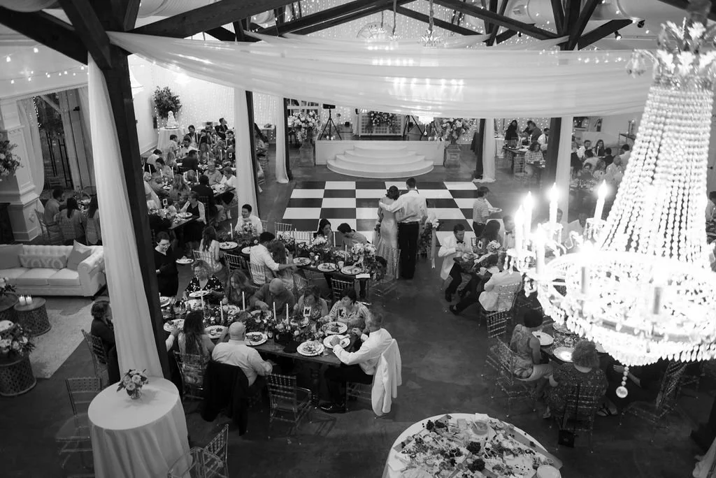 A large wedding and event banquet hall in Pensacola with round and rectangular tables decorated for a celebration, with many people dining, a dance floor, a stage, and a large crystal chandelier overhead.
