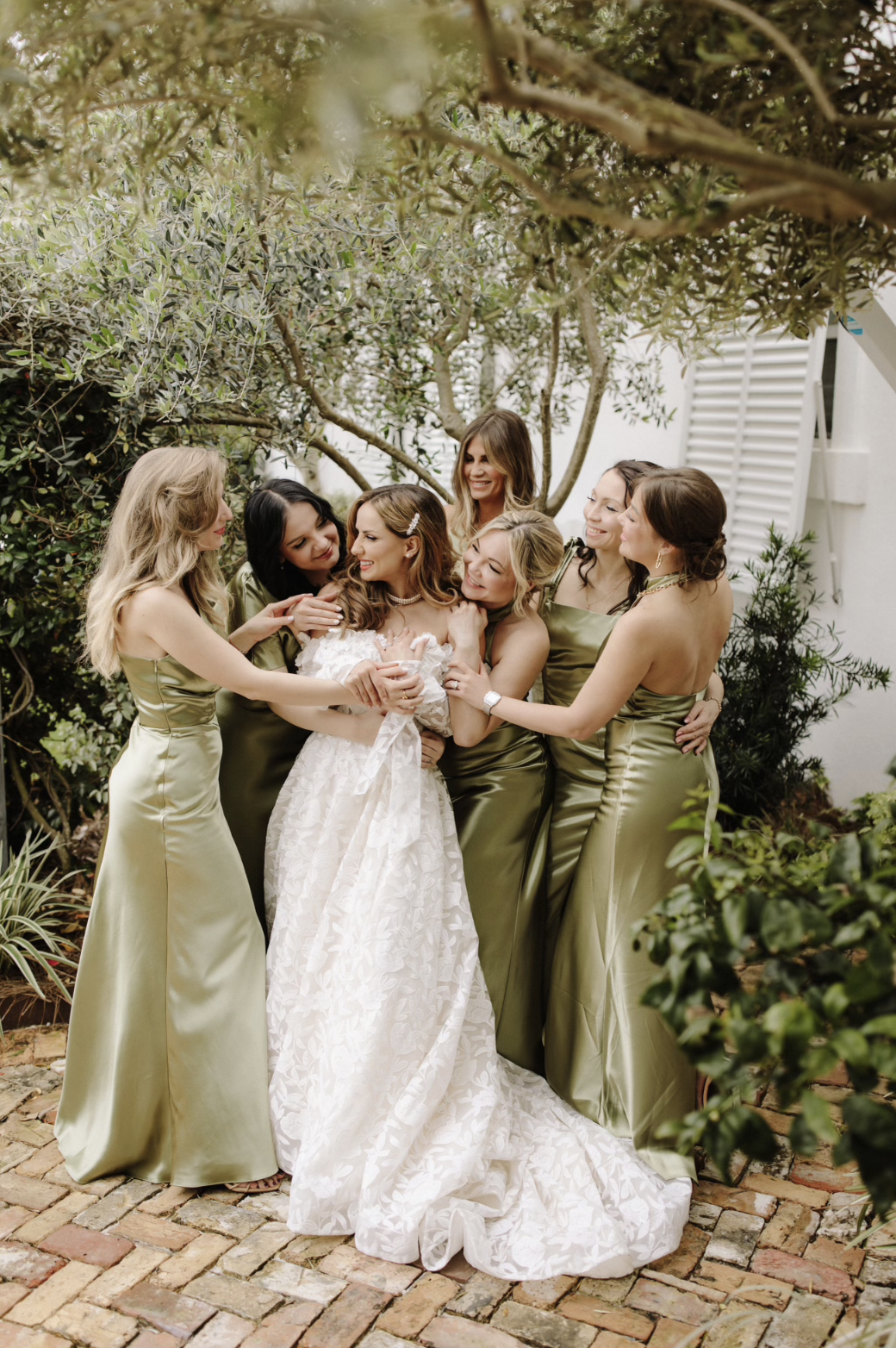 Bridesmaids in olive green satin dresses surrounding a bride in a white lace wedding gown, all smiling and embracing outdoors among trees and plants at Supposey.
