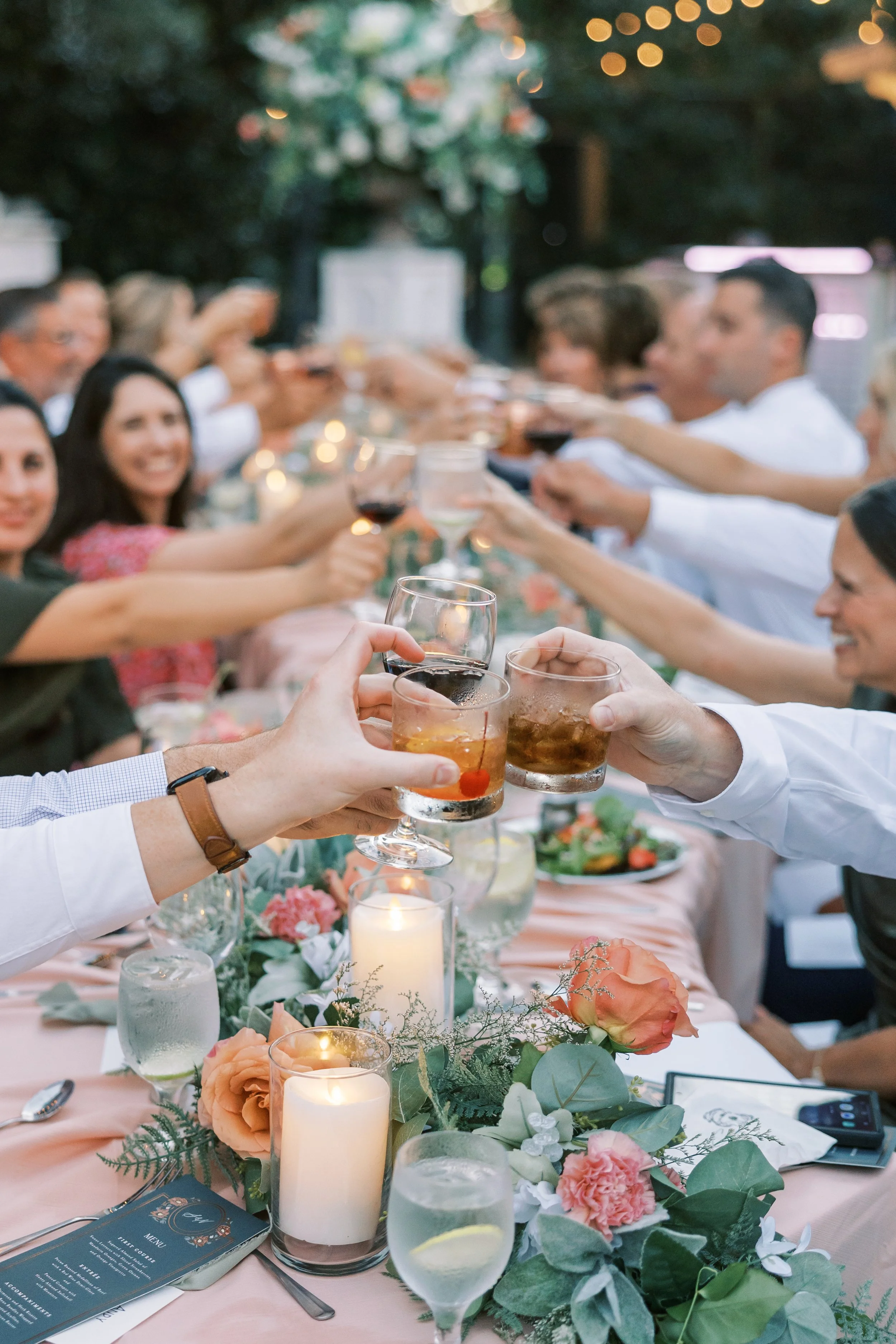 A group of people raising glasses in a toast at a dinner party outdoors with a long table decorated with flowers and candles in a Pensacola Wedding Venue.