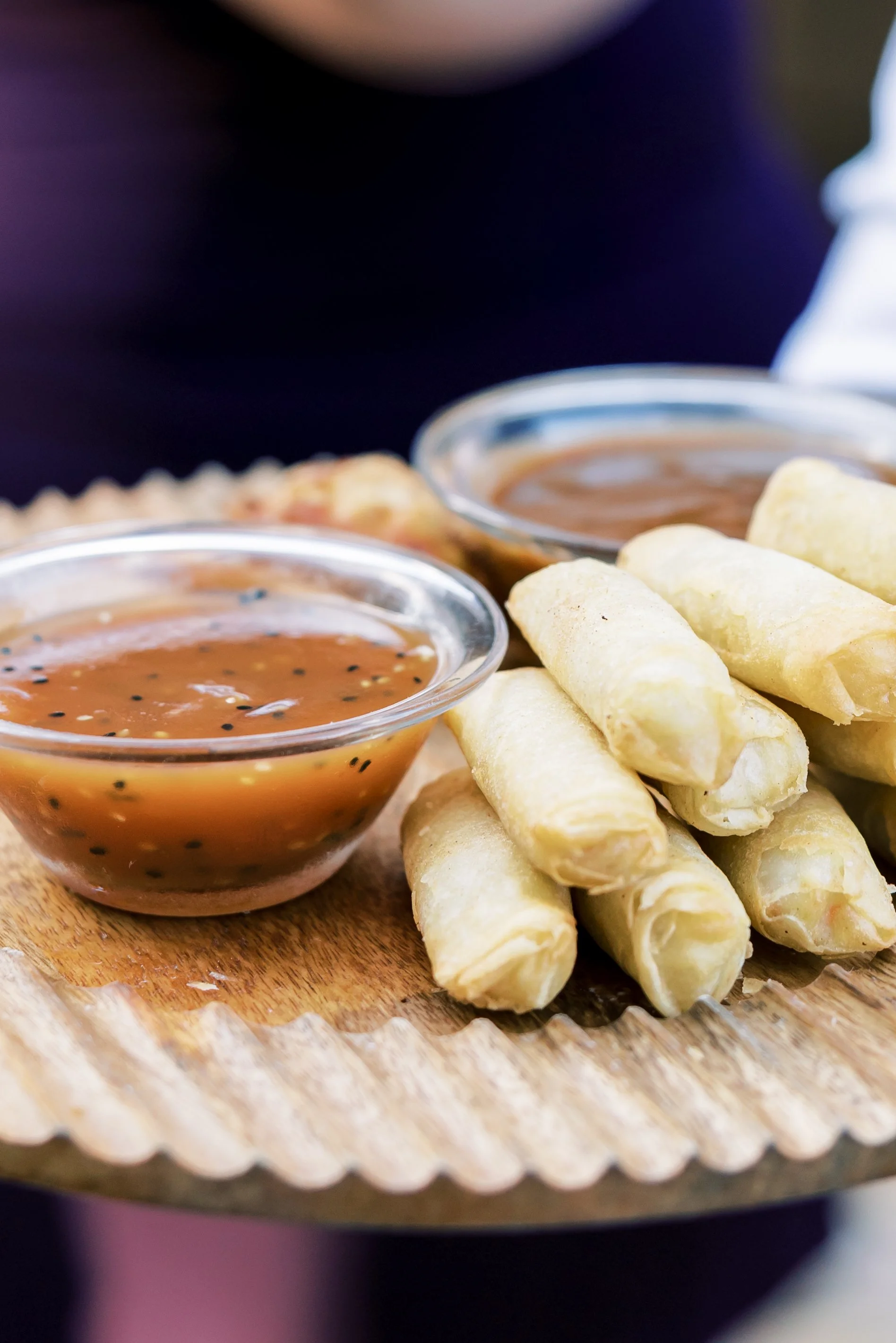 A wooden board with a pile of fried spring rolls and two small bowls of dipping sauce, one with sweet chili sauce and the other with brown sauce.