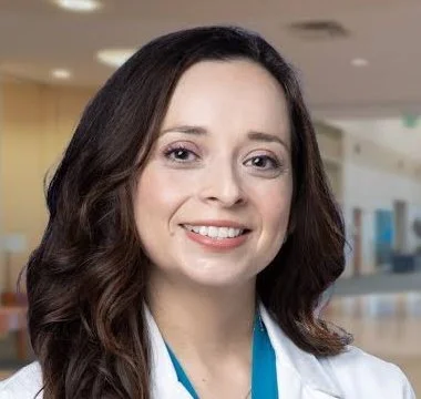 A woman with long brown hair smiling in a medical or hospital setting.