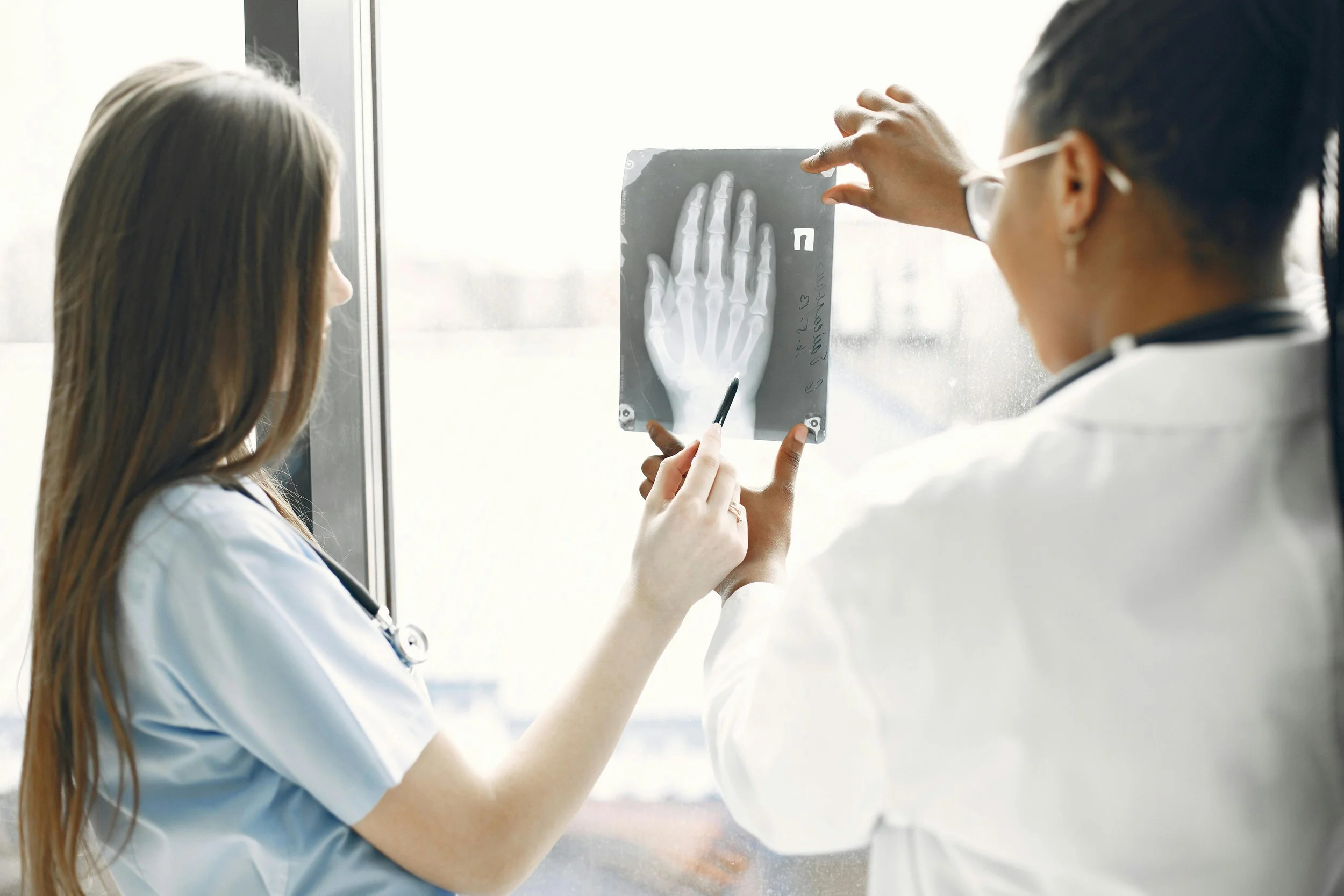 A female healthcare professional in a light blue uniform and a male healthcare professional in a white coat examining an X-ray of a hand.