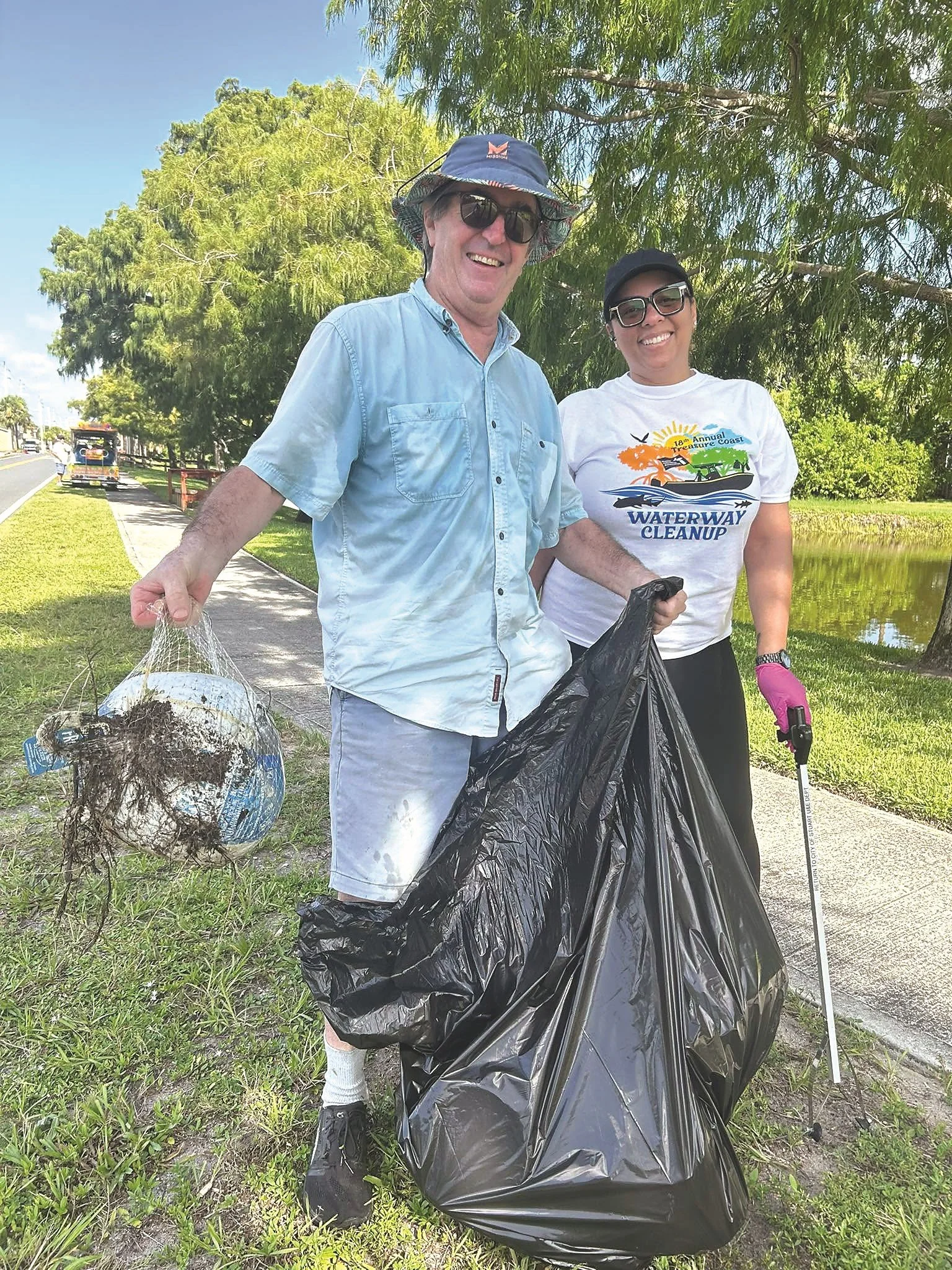 Commissioner Campbell Rich works with volunteers to clean up Treasure Coast waterways during Waterway Cleanup Day. With a big smile, he is joyfully holding a large black trash bag in one hand and some netting and debris in his other hand.