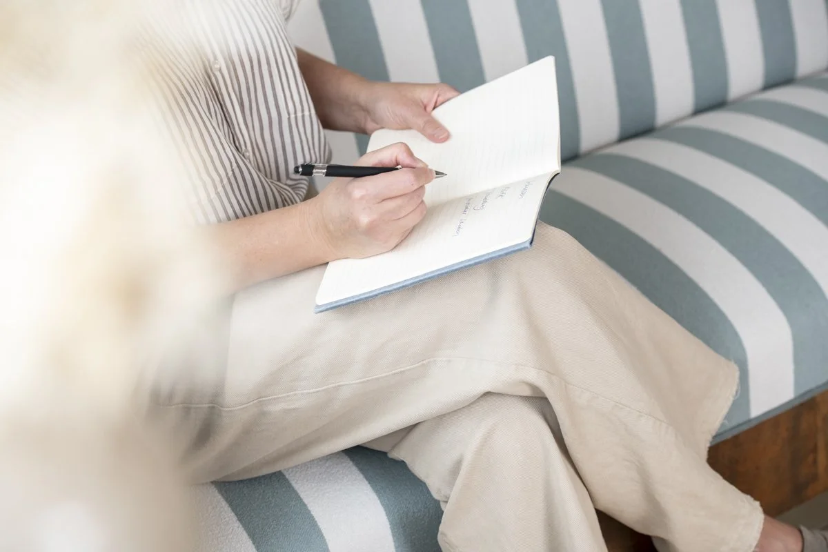 Person schreibt in ein Notizbuch, sitzt auf einem gepolsterten Sitz mit blauen und weißen Streifen, trägt beiges Kleid. Nahaufnahme, Fokus auf Hände und Notizbuch.