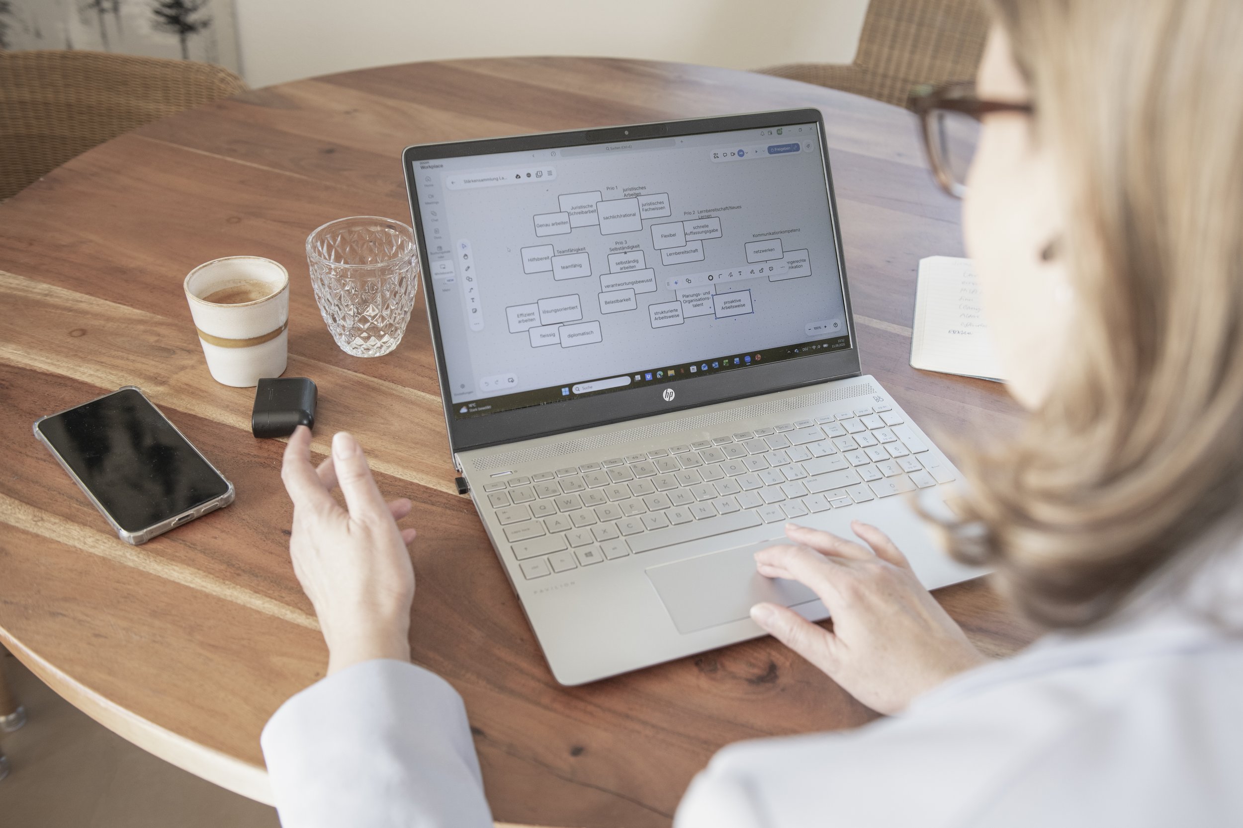 Frau arbeitet an einem Laptop auf einem Holztisch, es ist eine Notizbuch, ein Glas mit Wasser, eine Tasse Kaffee, ein Smartphone und ein Handy-Ladegerät auf dem Tisch.