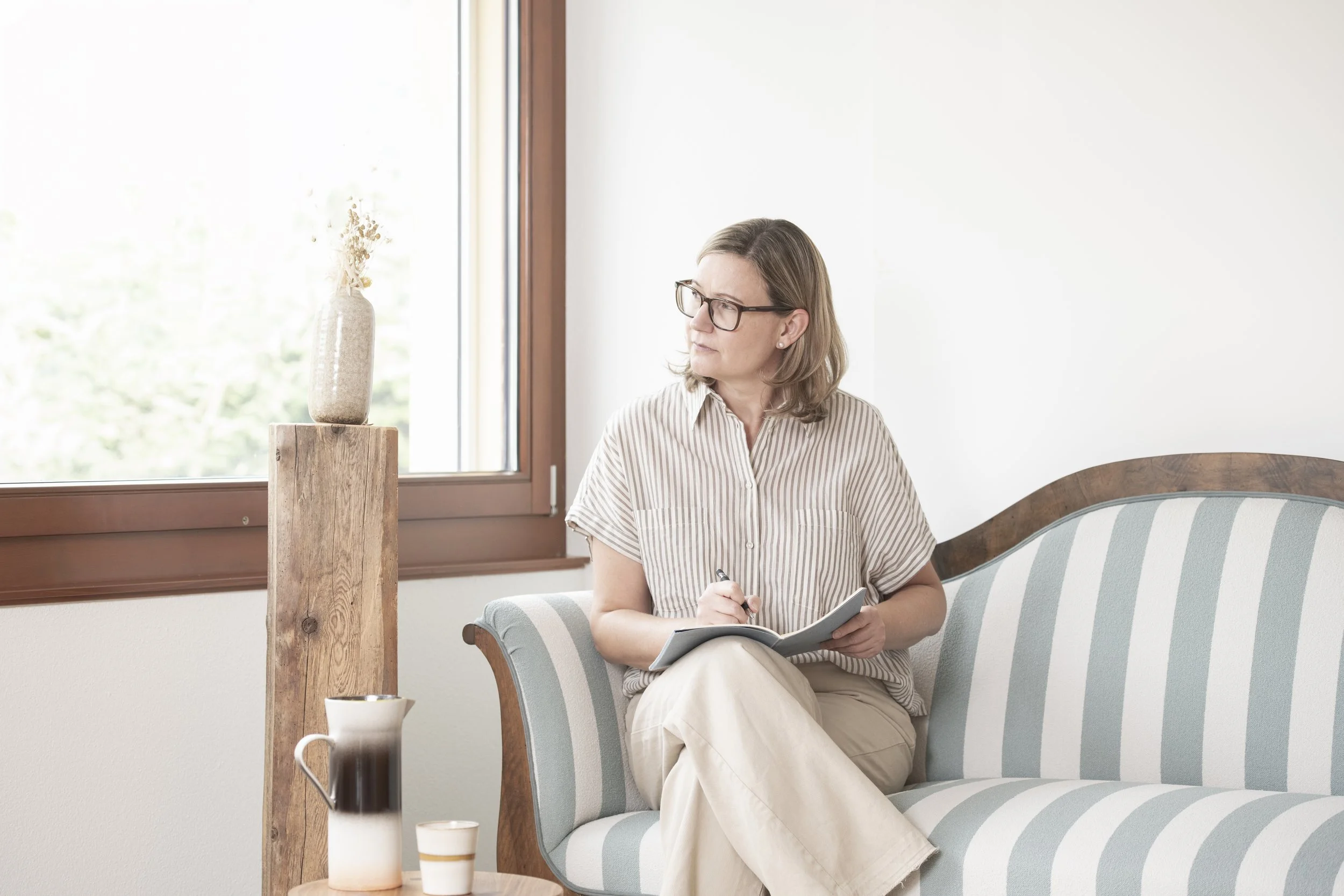 Eine Frau mit Brille sitzt auf einem Stuhl in einem hellen Raum, schreibt in ein Notizbuch, neben ihr ein Tisch mit einer Kaffeetasse und einem Flüssigkeitsbehälter, im Hintergrund ein Fenster mit Blick nach draußen.