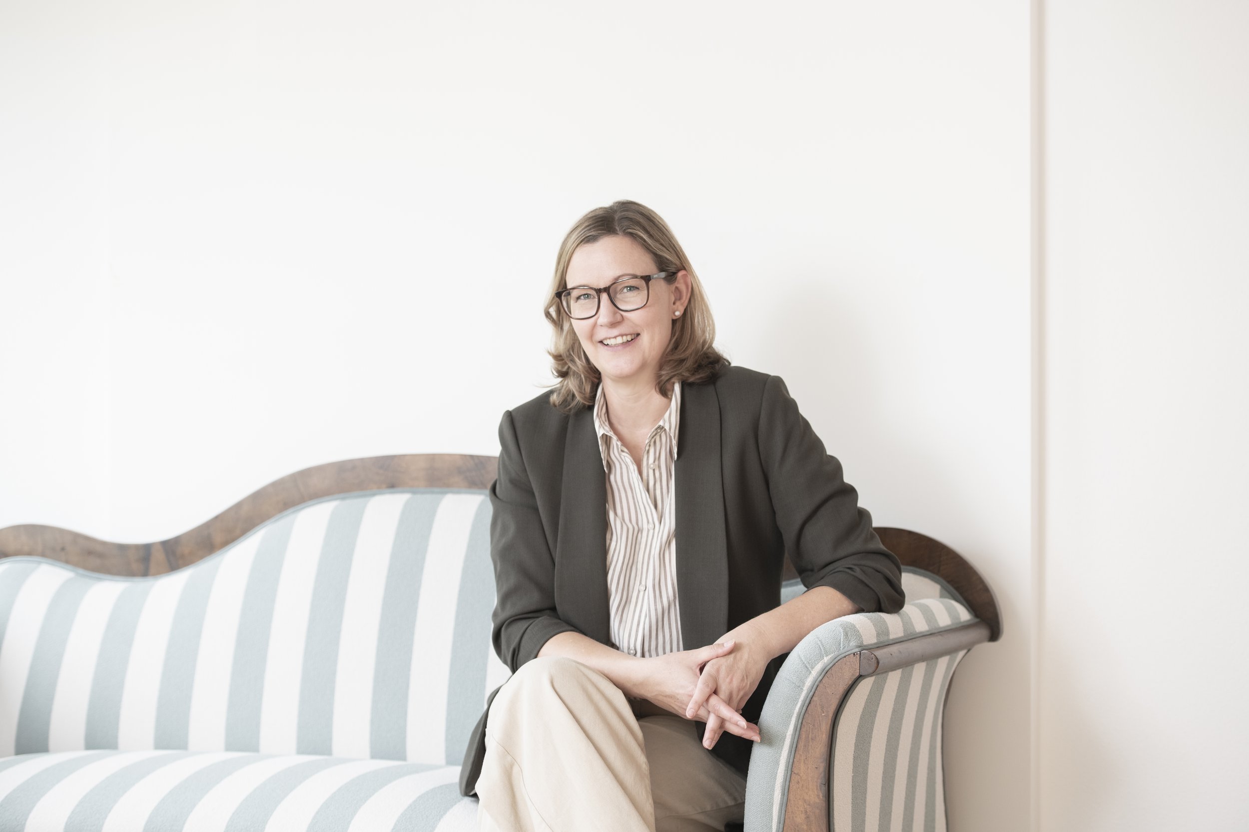 Eine lachende Frau mit Brille, die in einem schwarzen Blazer und einer gestreiften Bluse sitzt, auf einem gepolsterten Sitzen im Wartezimmer oder Büro gegenüber einem weißen Wand. Sie hat mittellange braune Haare und wirkt professionell und freundlich.