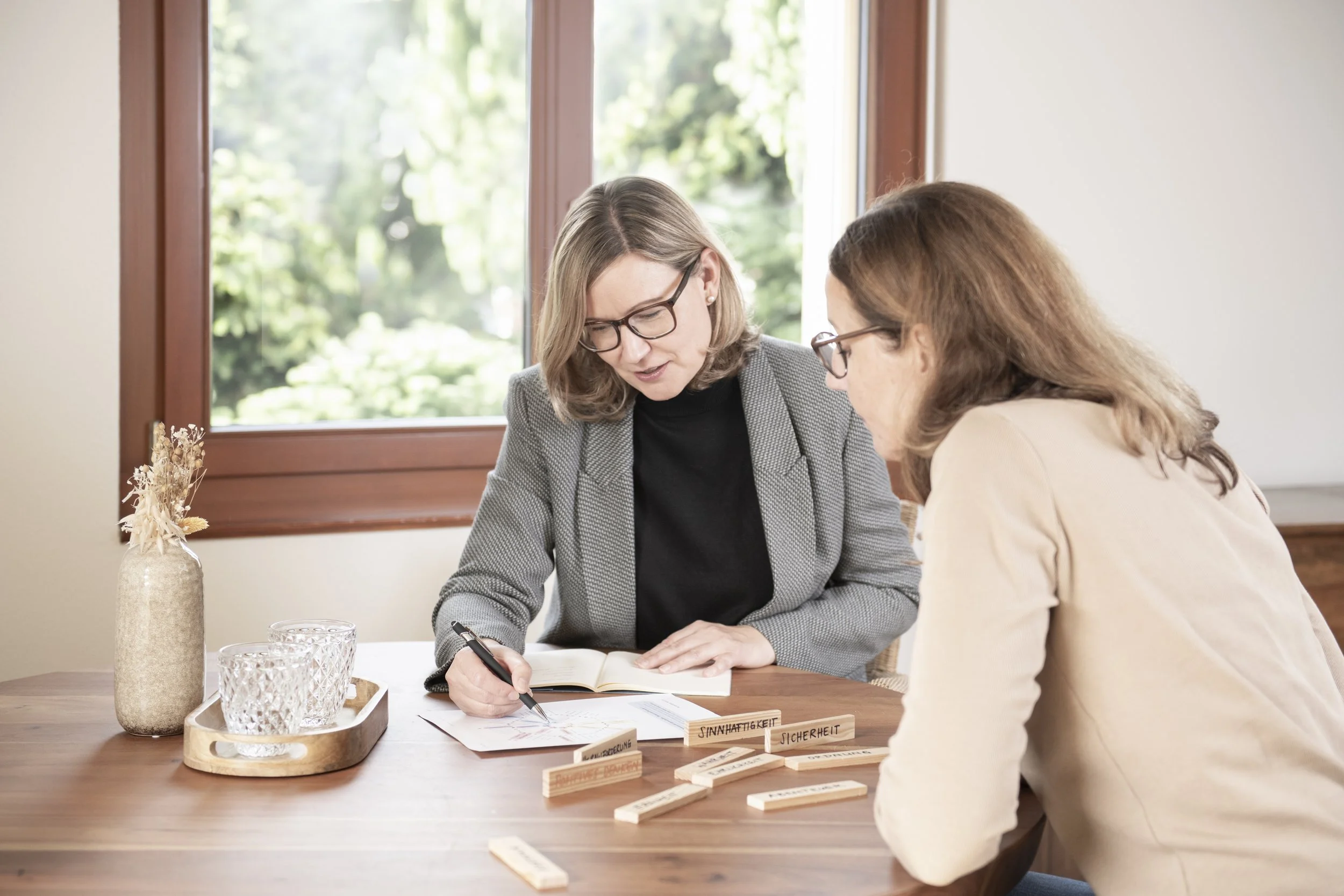 Zwei Frauen sitzen an einem Holztisch, eine schreibt in ein Notizbuch, die andere schaut zu. Im Vordergrund befinden sich Holzklötze mit deutschen Wörtern wie "Sinnhaftigkeit", "Sicherheit" und "Zuverlässigkeit".