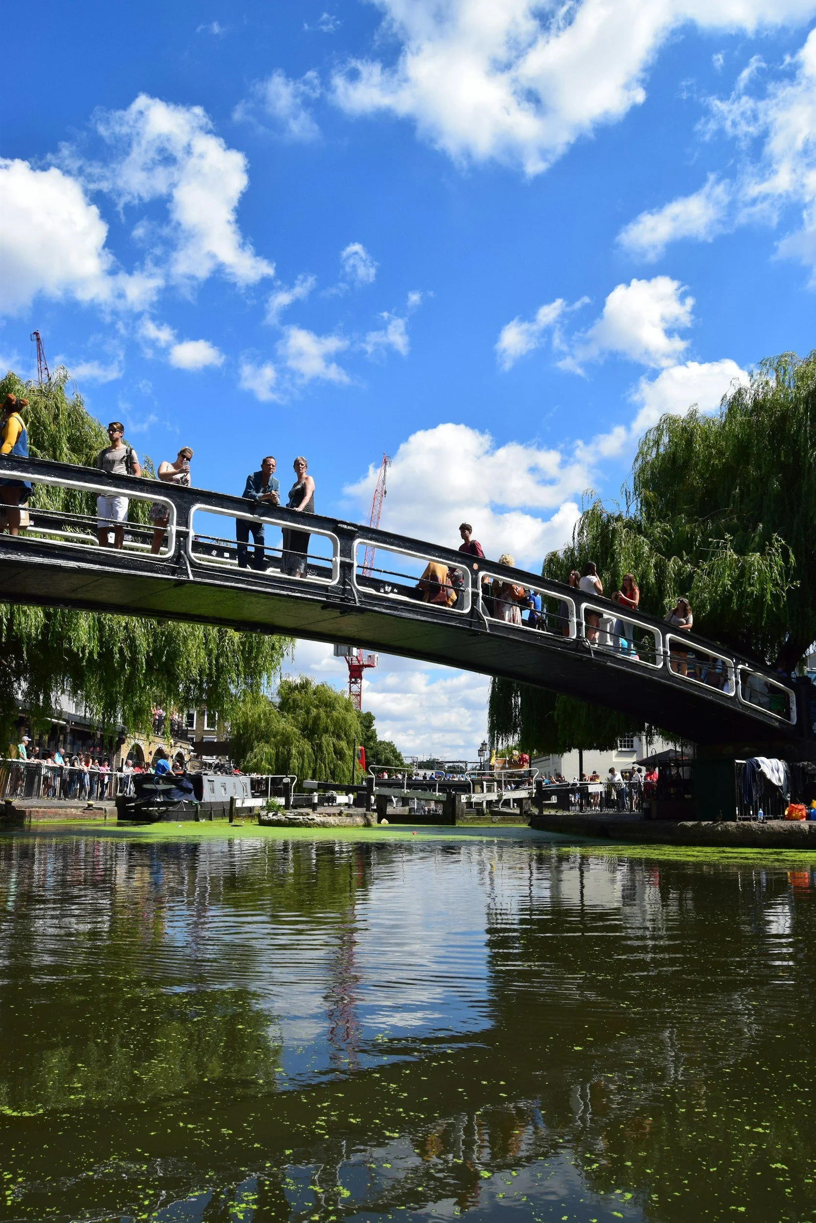 Central Victorian House | Camden Town | People walking on a footbridge over a canal on a sunny day with blue sky and clouds. Trees line the canal banks, and reflections can be seen in the water.