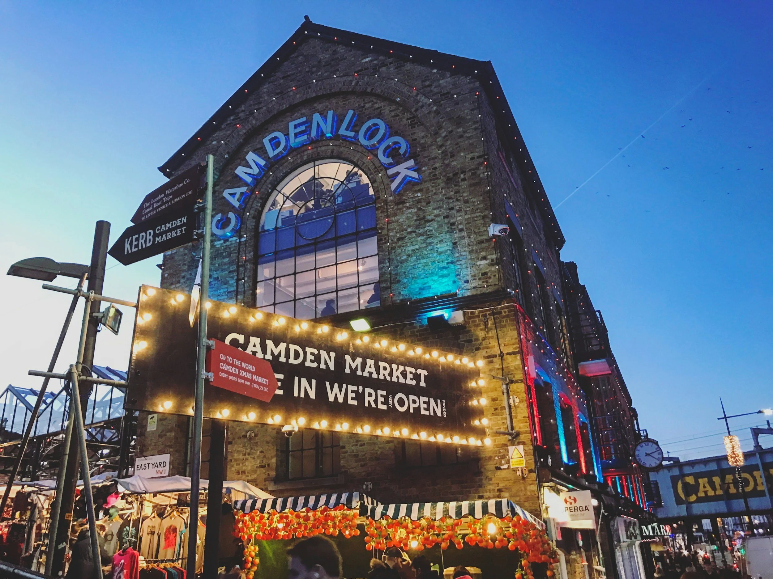 Central Victorian House | Camden Town | View of Camden Market in London during evening with illuminated signs, a brick building, and market stalls with umbrellas.
