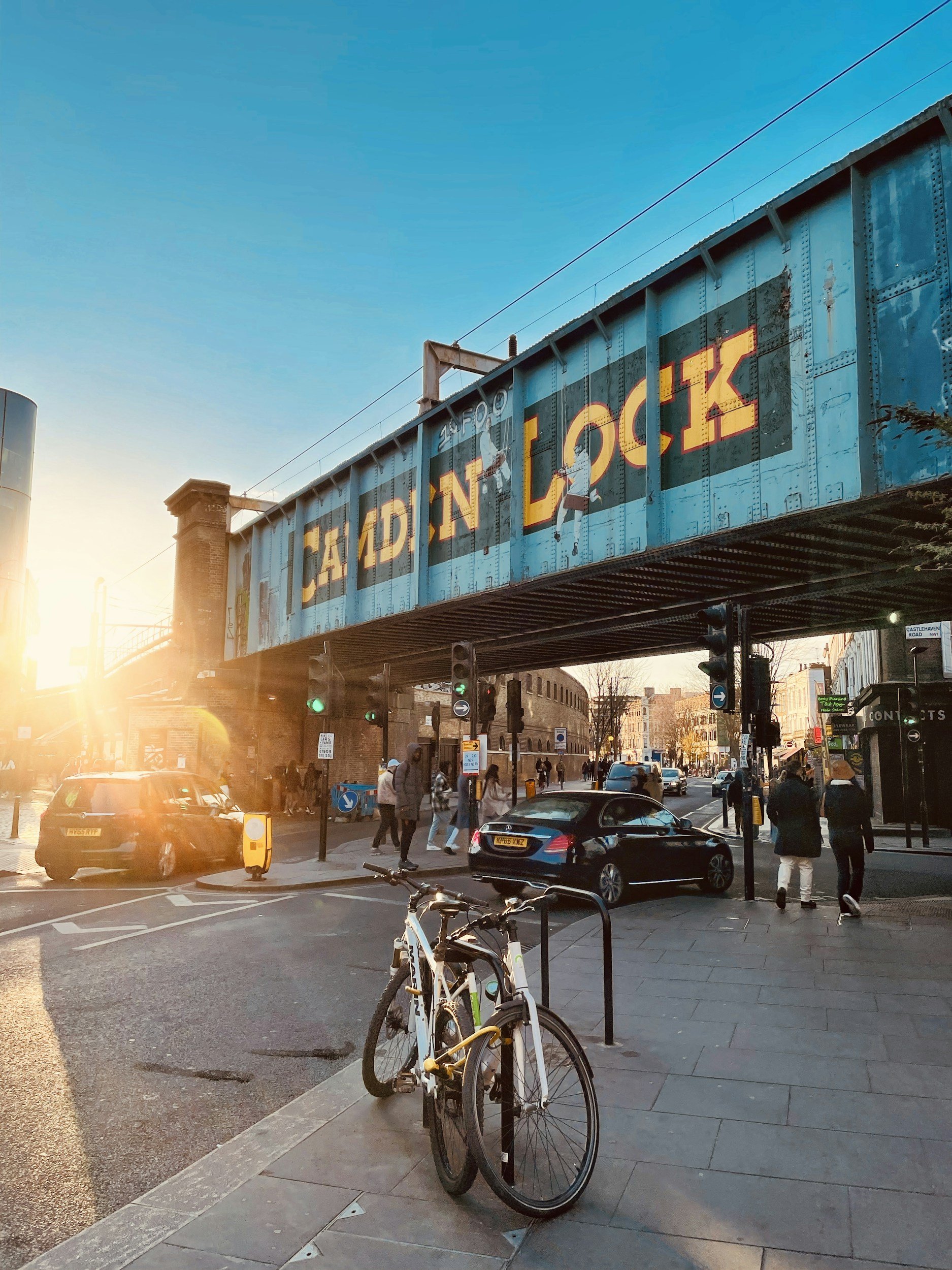 Central Victorian House | The Hawley | Camden Lock in Camden Town | A city street scene during sunset with a cyclist's bicycle parked on the sidewalk, cars on the road, pedestrians walking, and a railway bridge with painted text and artwork overhead.