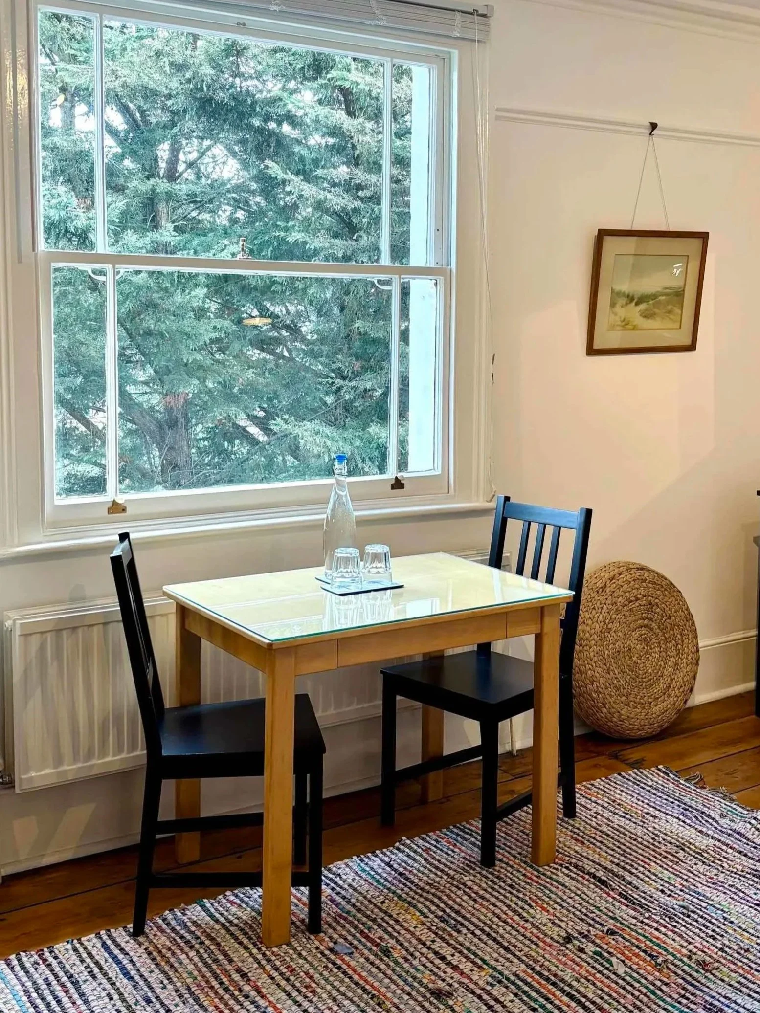 A dining room with a wooden table, two black chairs, a colorful woven rug, windows showing trees outside, and a wicker object next to the wall. There is a bottle and three glasses on the table, and a framed landscape picture on the wall.