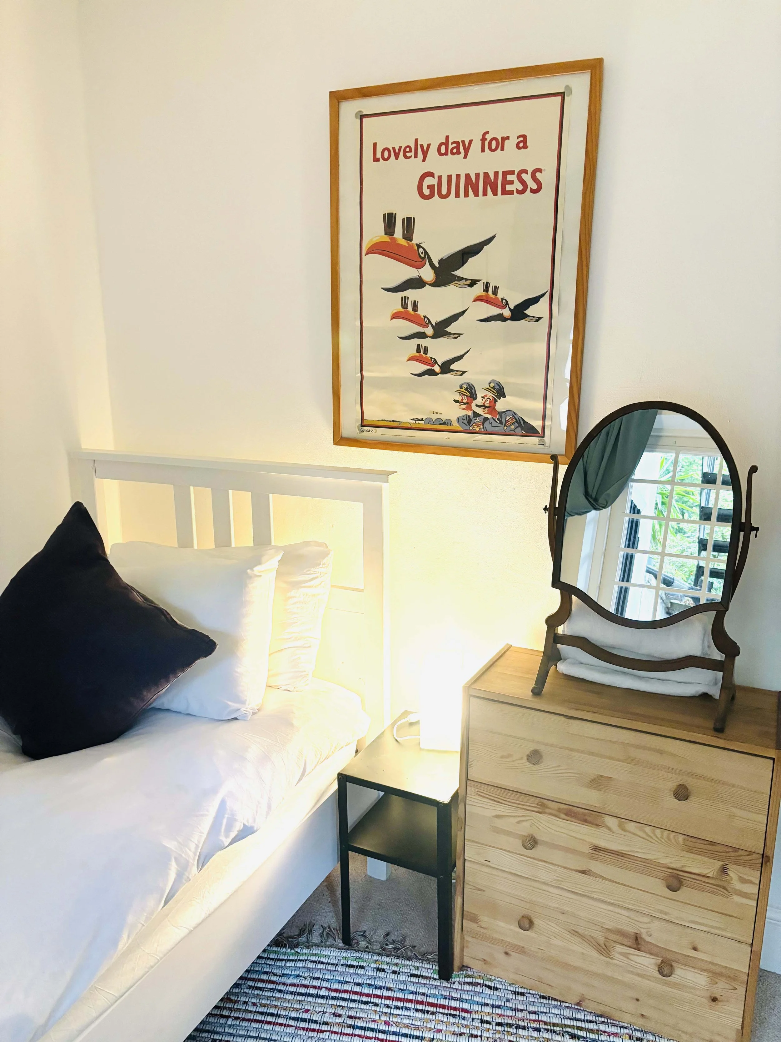 Central Victorian House | A bedroom corner with a bed, pillows, a dresser with a mirror, a small black table, and a wall poster featuring a toucan with the text 'Lovely day for a Guinness'.